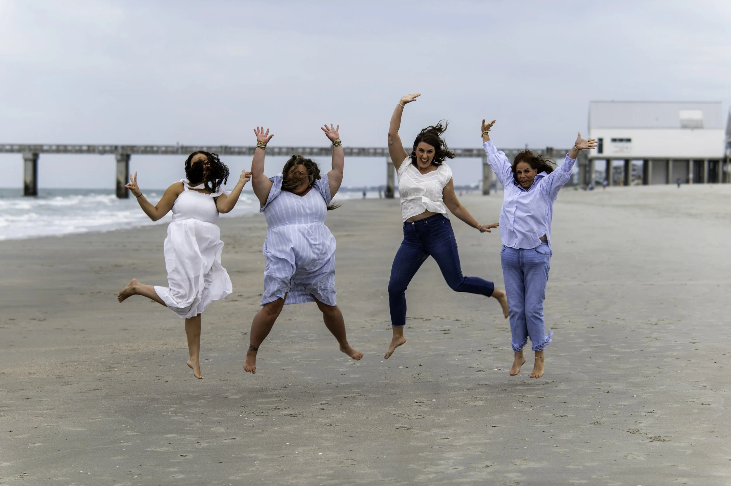 Four women are jumping and smiling on a beach with a pier in the background. They appear to be enjoying a fun moment together.