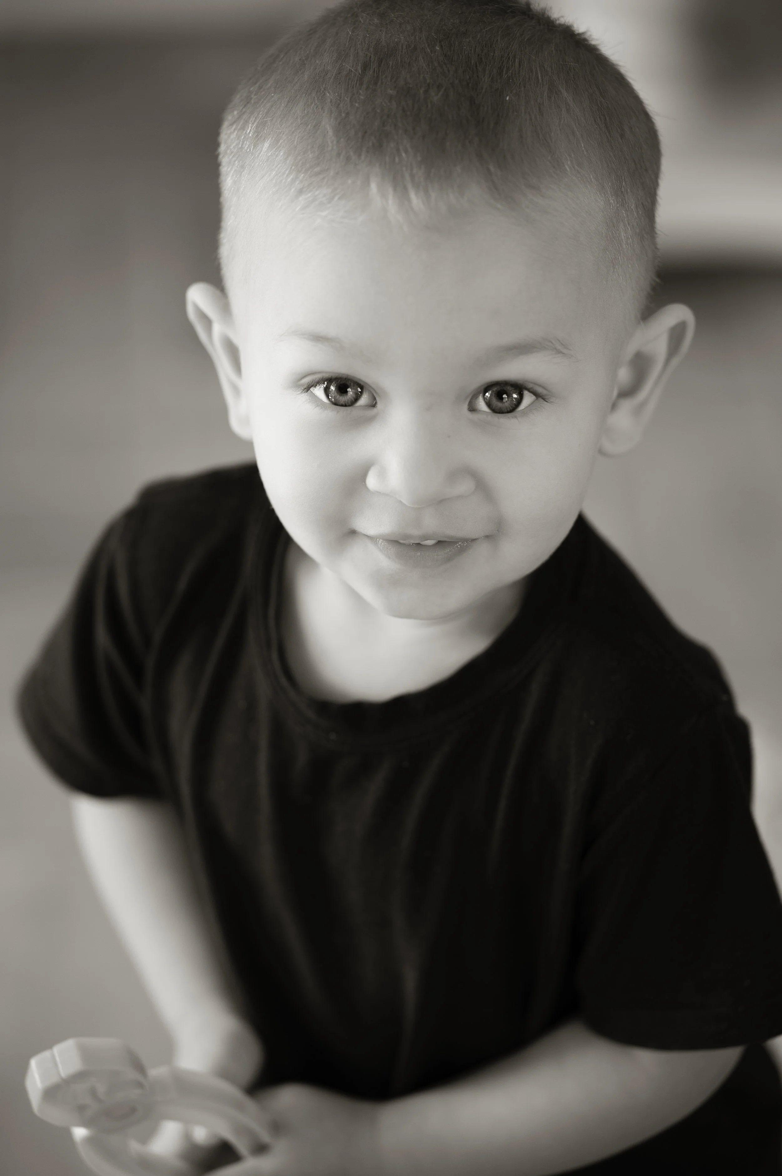 A young boy with short hair and light-colored eyes, wearing a black t-shirt, smiling and looking up at the camera, holding a toy in his hand.
