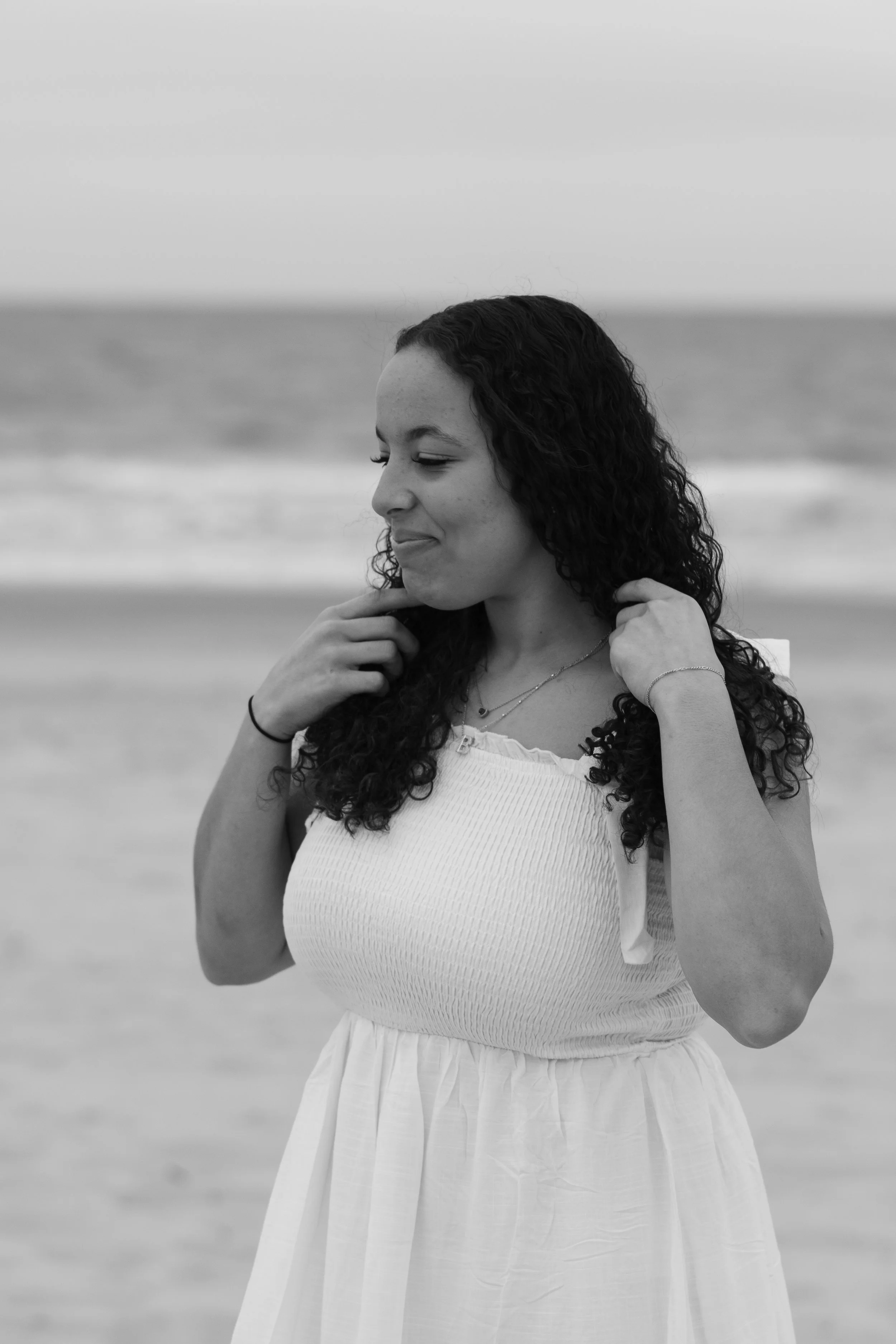 A young woman with long, curly hair stands on the beach with her eyes closed and a gentle smile. She is touching her shoulders with her hands, wearing a white textured dress and layered necklaces, with the ocean and sky blurred in the background.