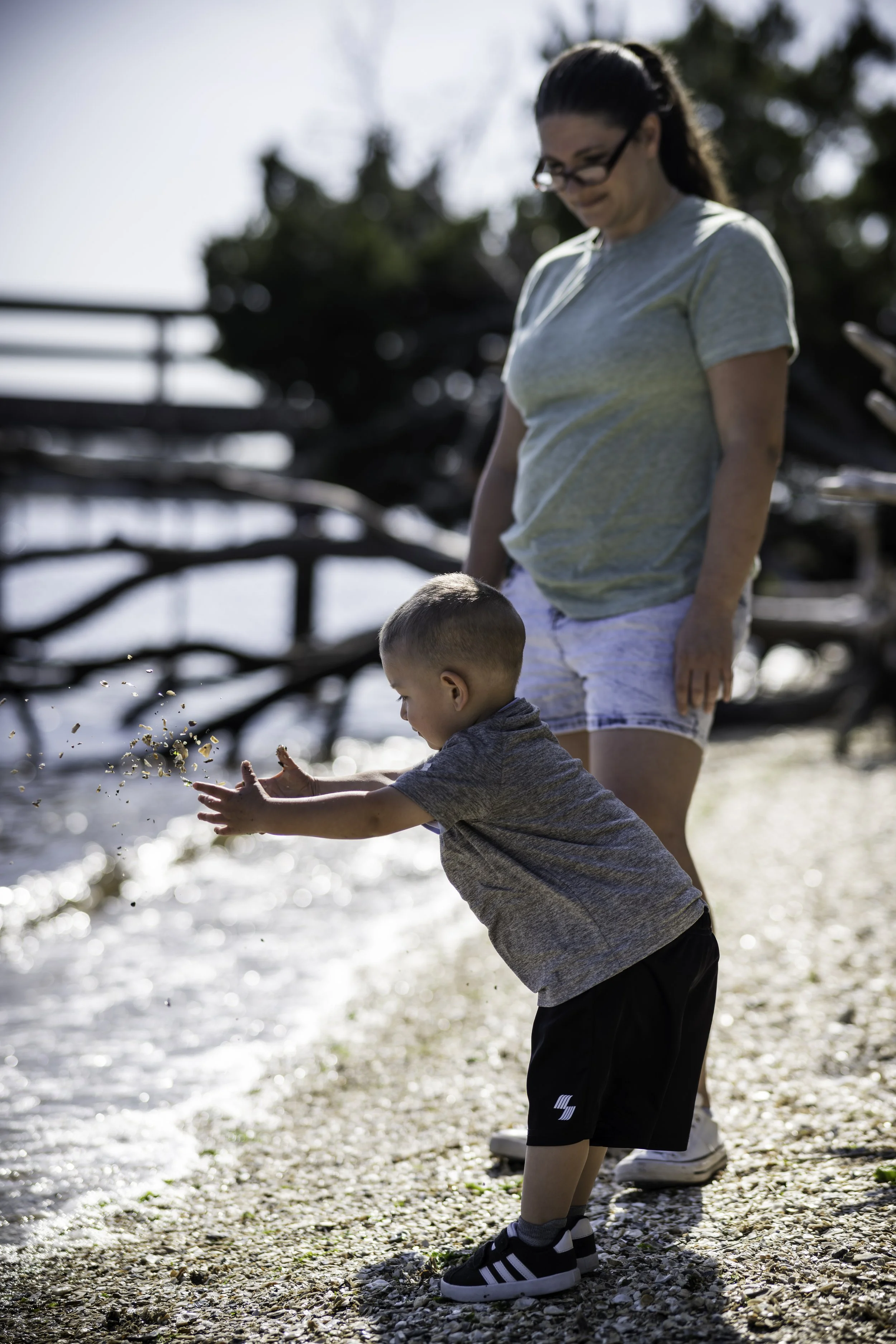 A woman and young boy at the beach, with the boy throwing sand by the water's edge.