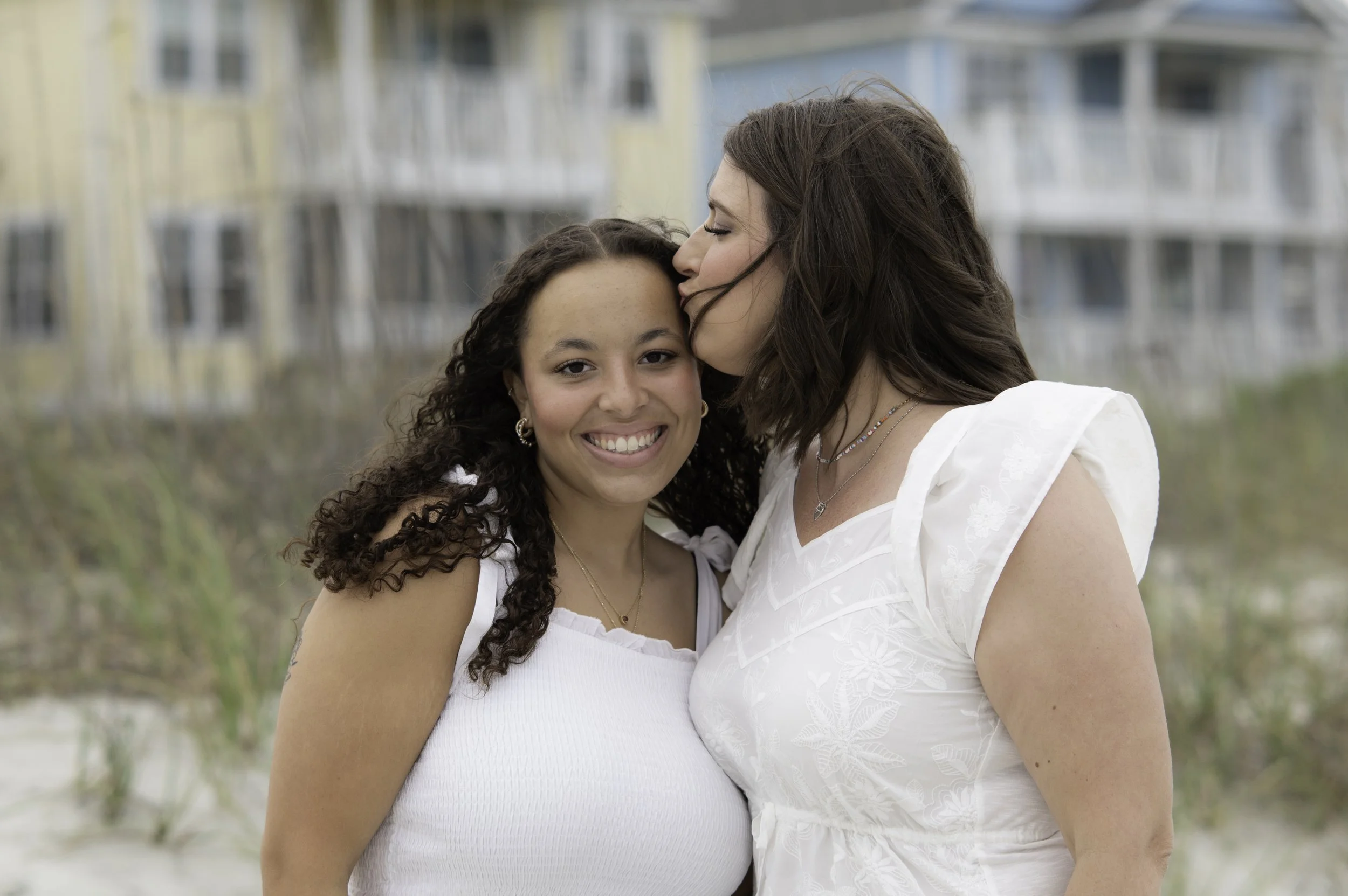 Two women, one kissing the other's forehead, standing outdoors with a background of blurred houses and greenery.