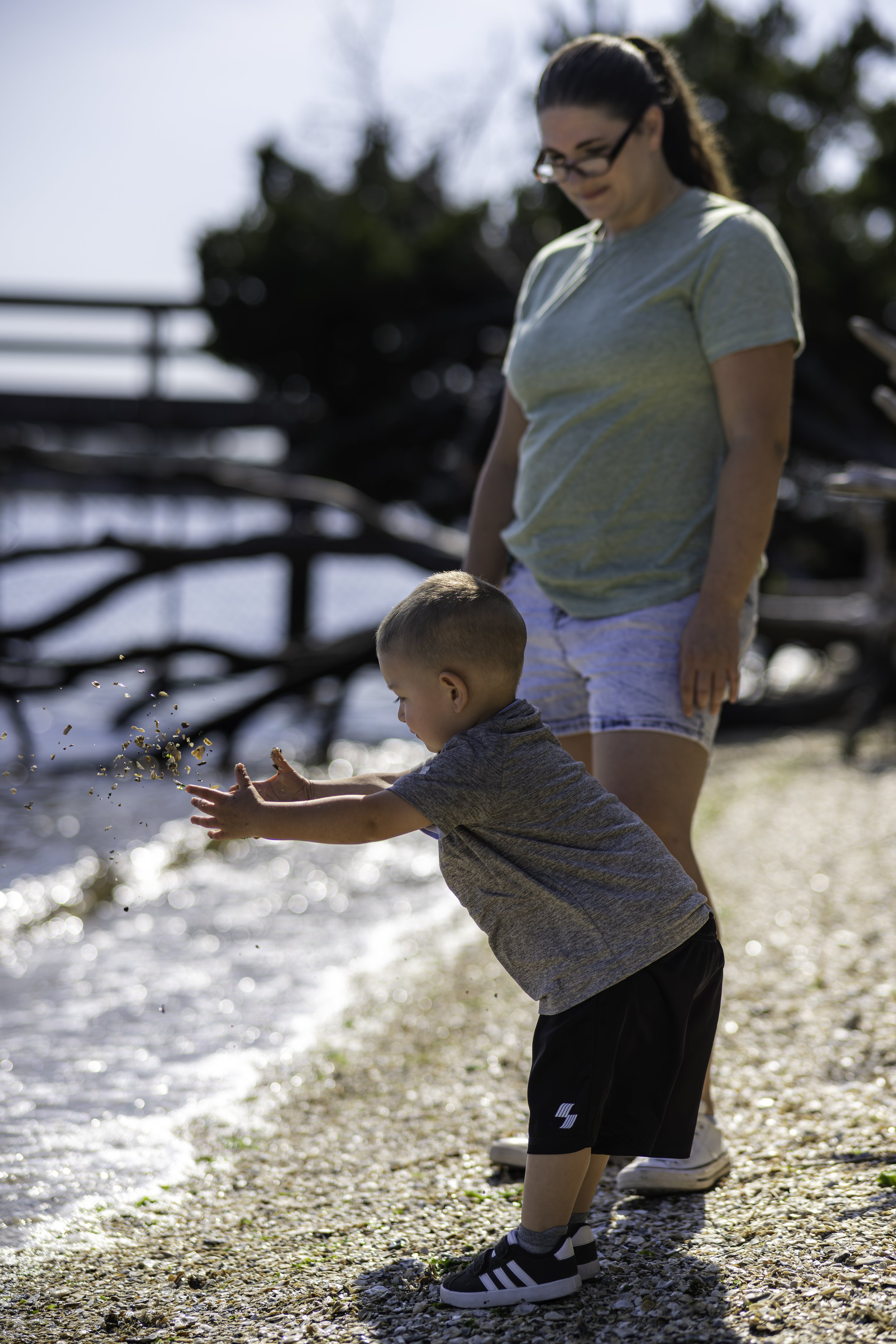 A woman and a young boy standing on a beach with shells, playing and splashing water.
