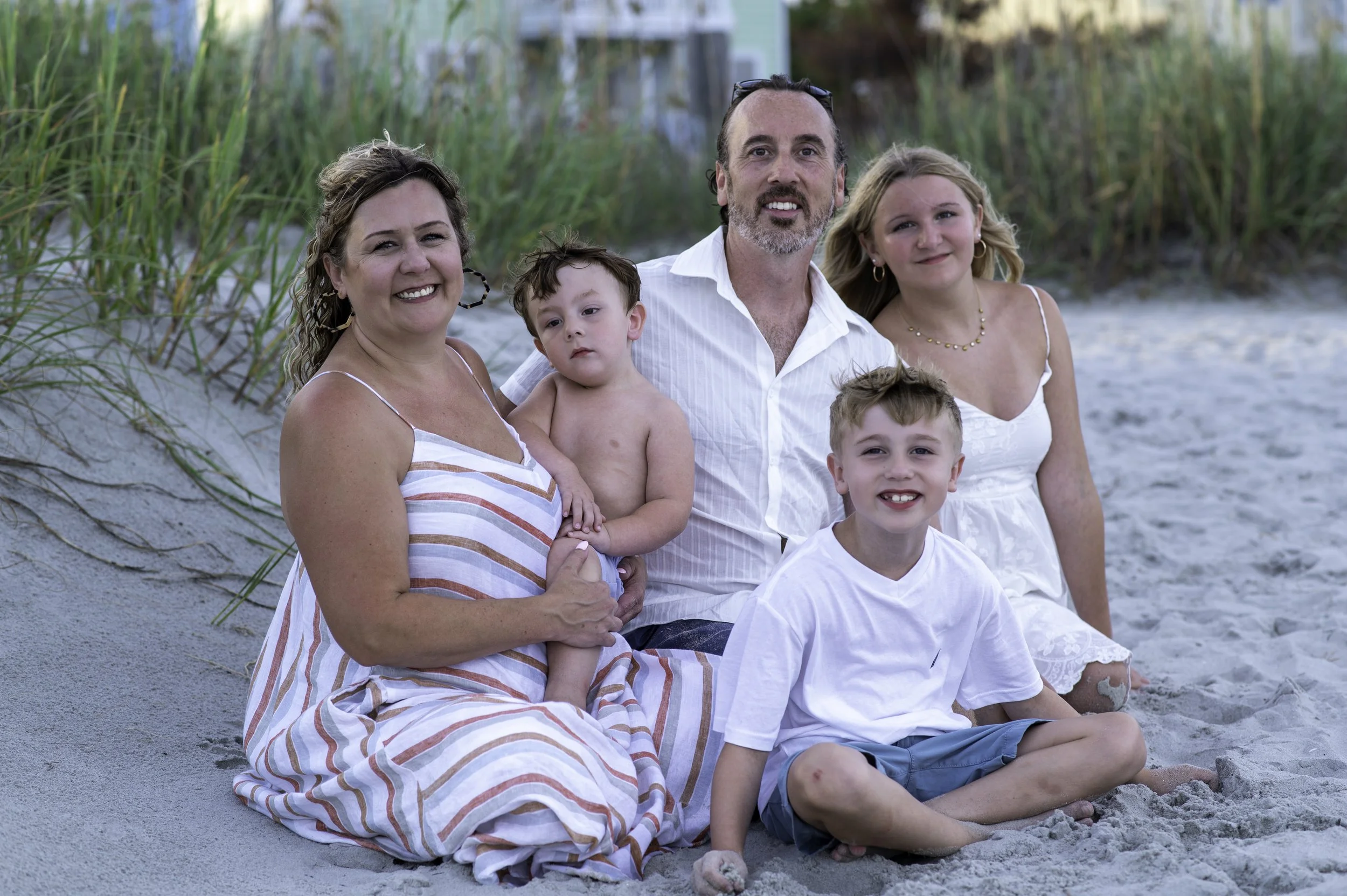 Family of five sitting on the sandy beach with tall grass in the background, smiling and enjoying a day outdoors.