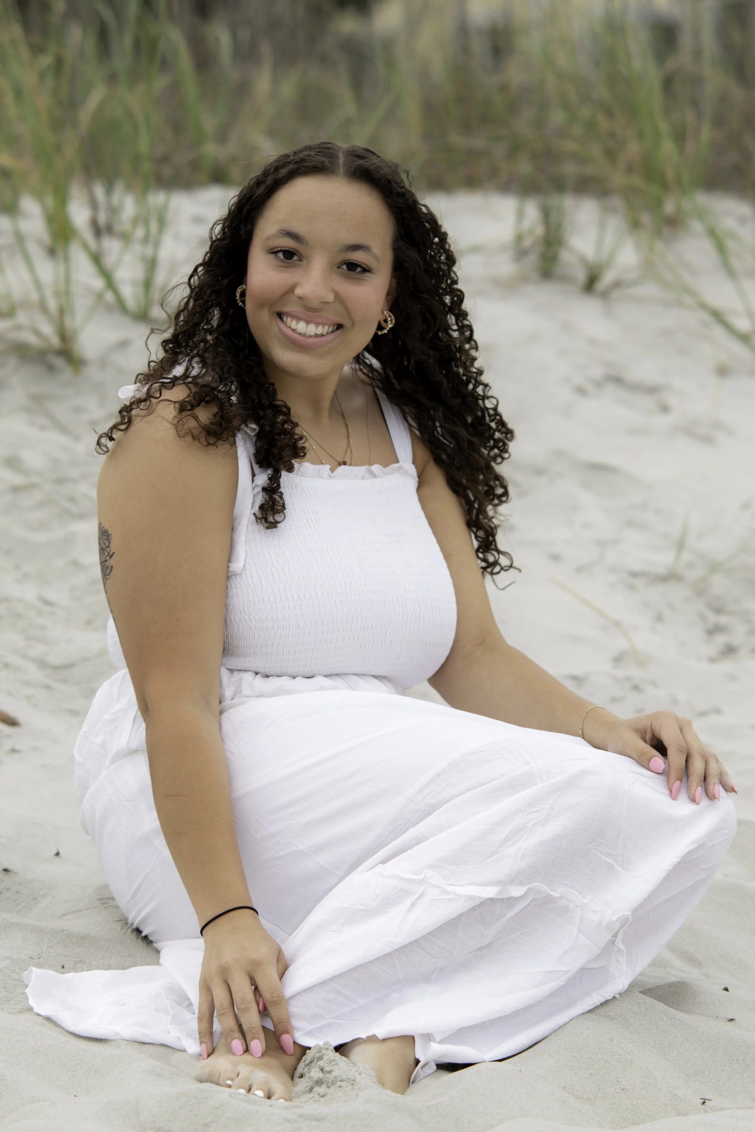 A young woman with curly hair, wearing a white tank top and white skirt, sitting on a sandy beach with green grasses in the background, smiling and looking at the camera.