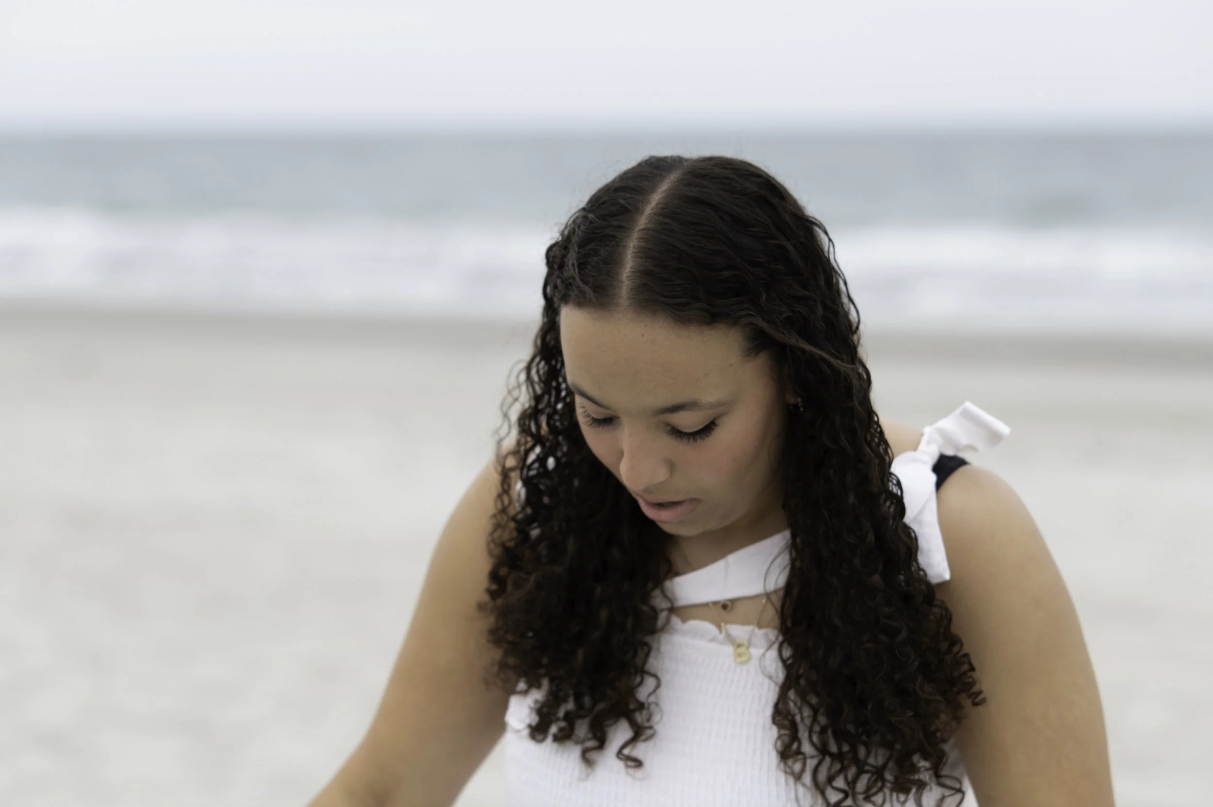 A young woman with long, curly dark hair looking down on a beach with a blurred ocean background.
