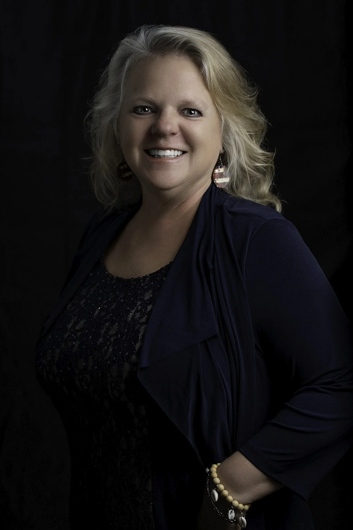 A smiling woman with blonde hair, wearing earrings, a dark top, a navy blue jacket, and a pearl bracelet, against a black background.
