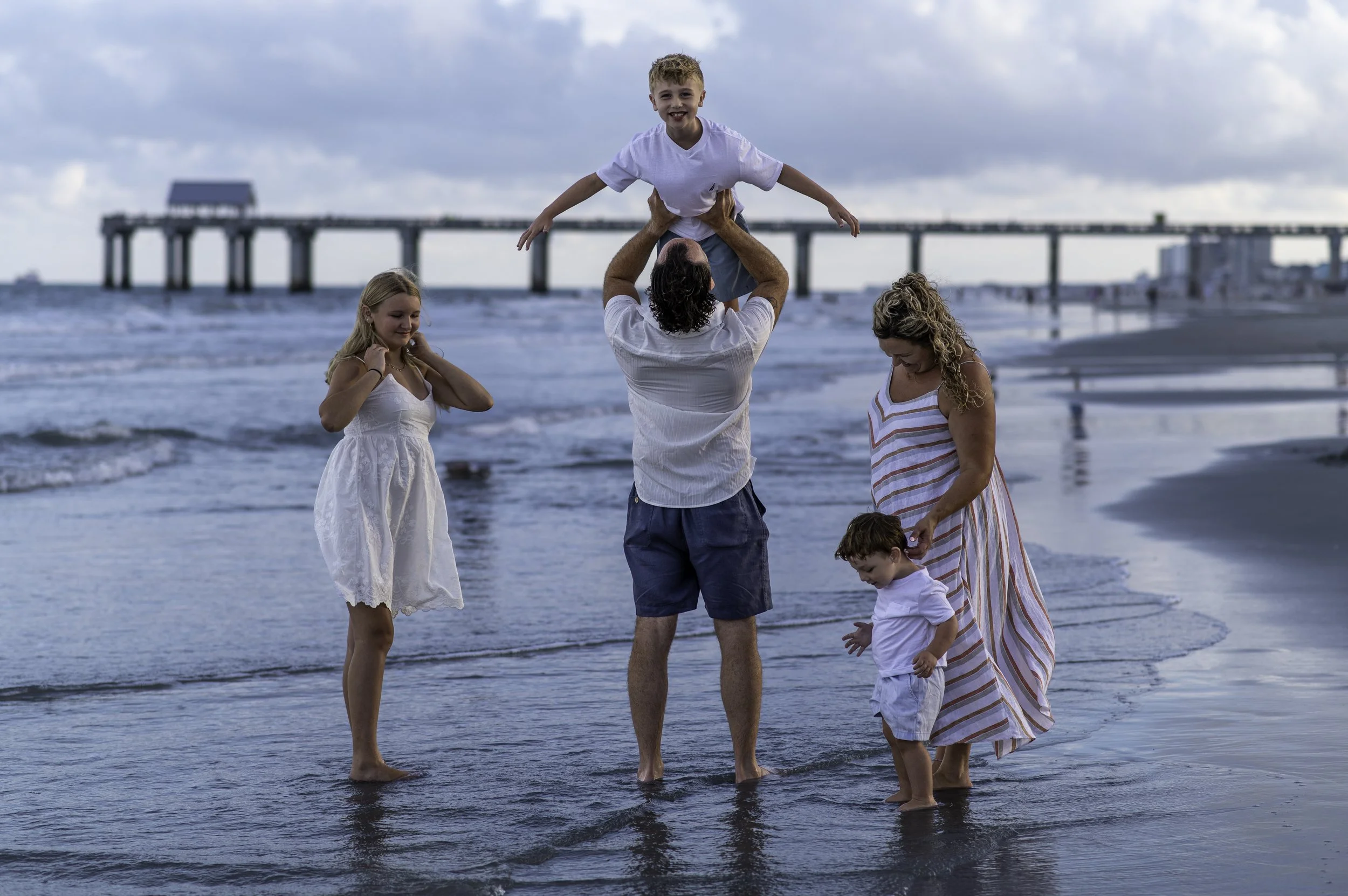 A group of five people, including two children, are standing on the beach at sunset, with a pier in the background. One child is being lifted in the air by an adult, while others are looking down at the water.