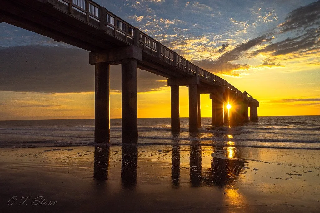 A pier extending over the ocean at sunset with the sun setting behind the pier's supports, casting a golden light on the water and sky.