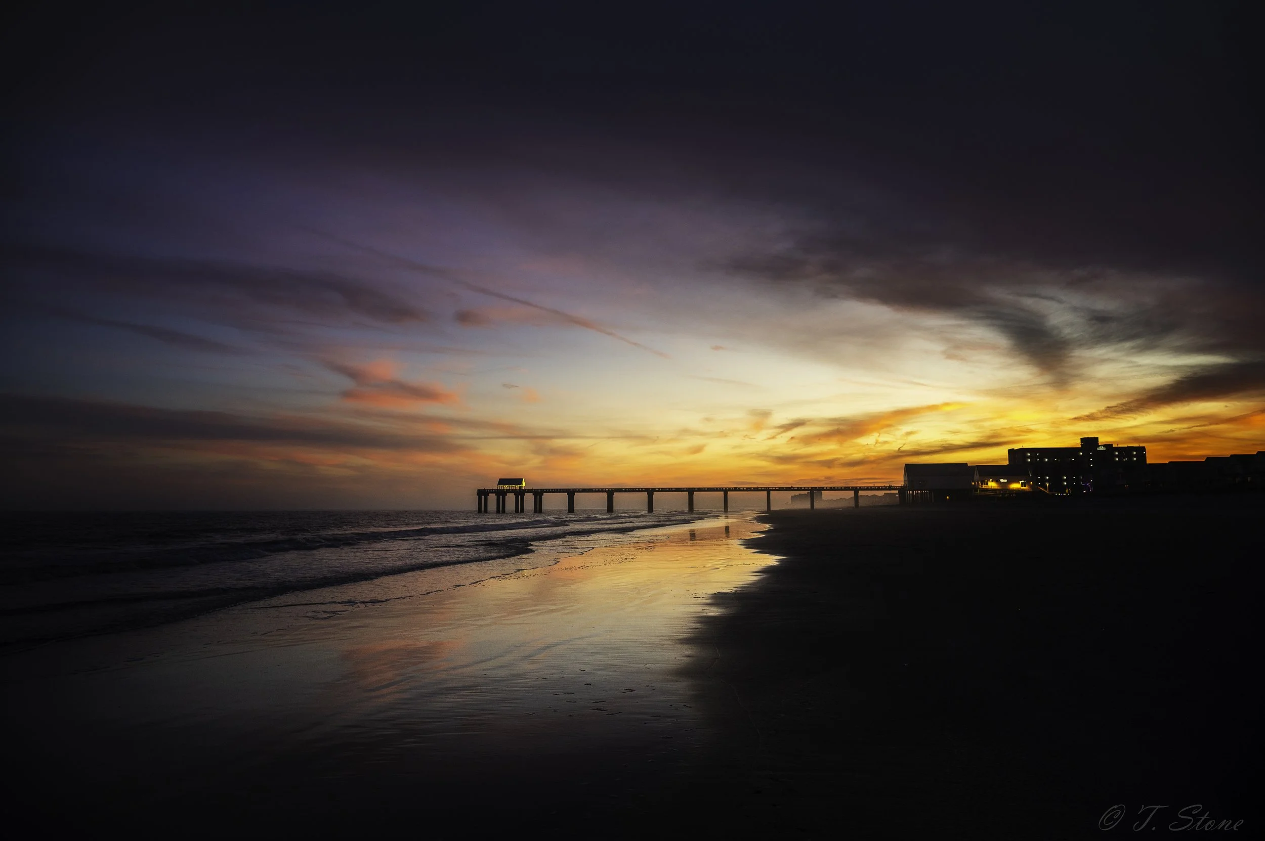 Sunset over a beach with a pier extending into the ocean and buildings in the distance, reflected in wet sand.