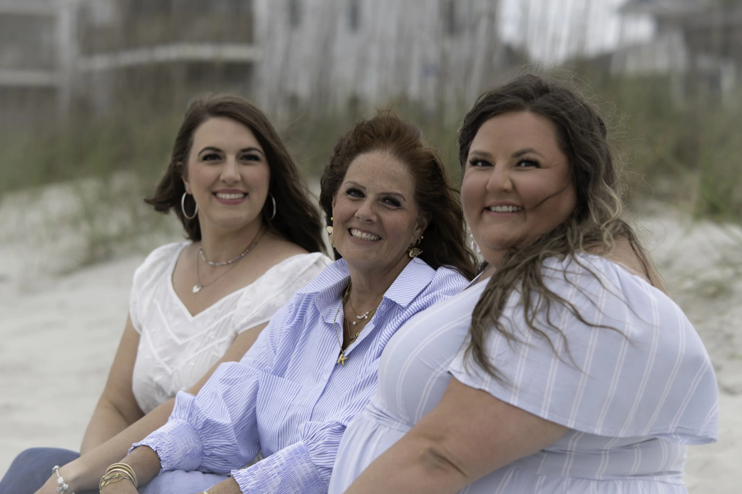 Four women sitting on a beach, smiling and posing for the photo. They are dressed in casual summer clothes.
