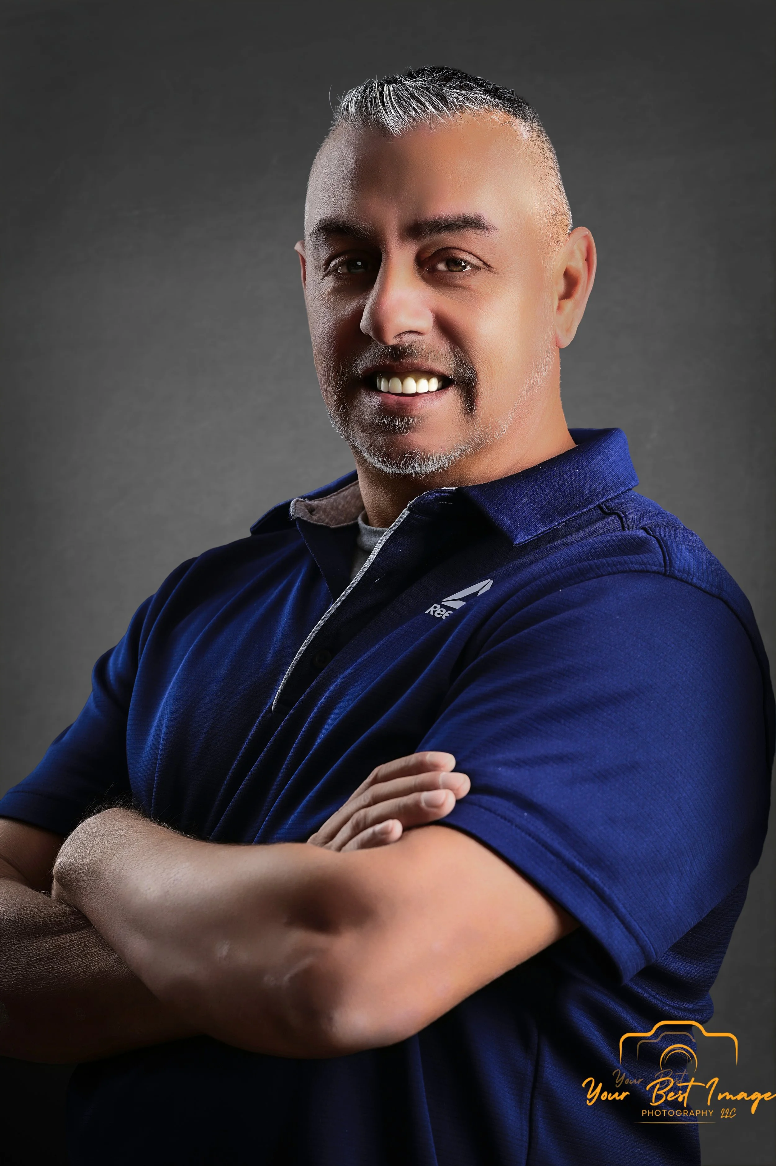 A smiling man with short, gray hair and a goatee, wearing a navy blue polo shirt, poses with arms crossed against a gray background.