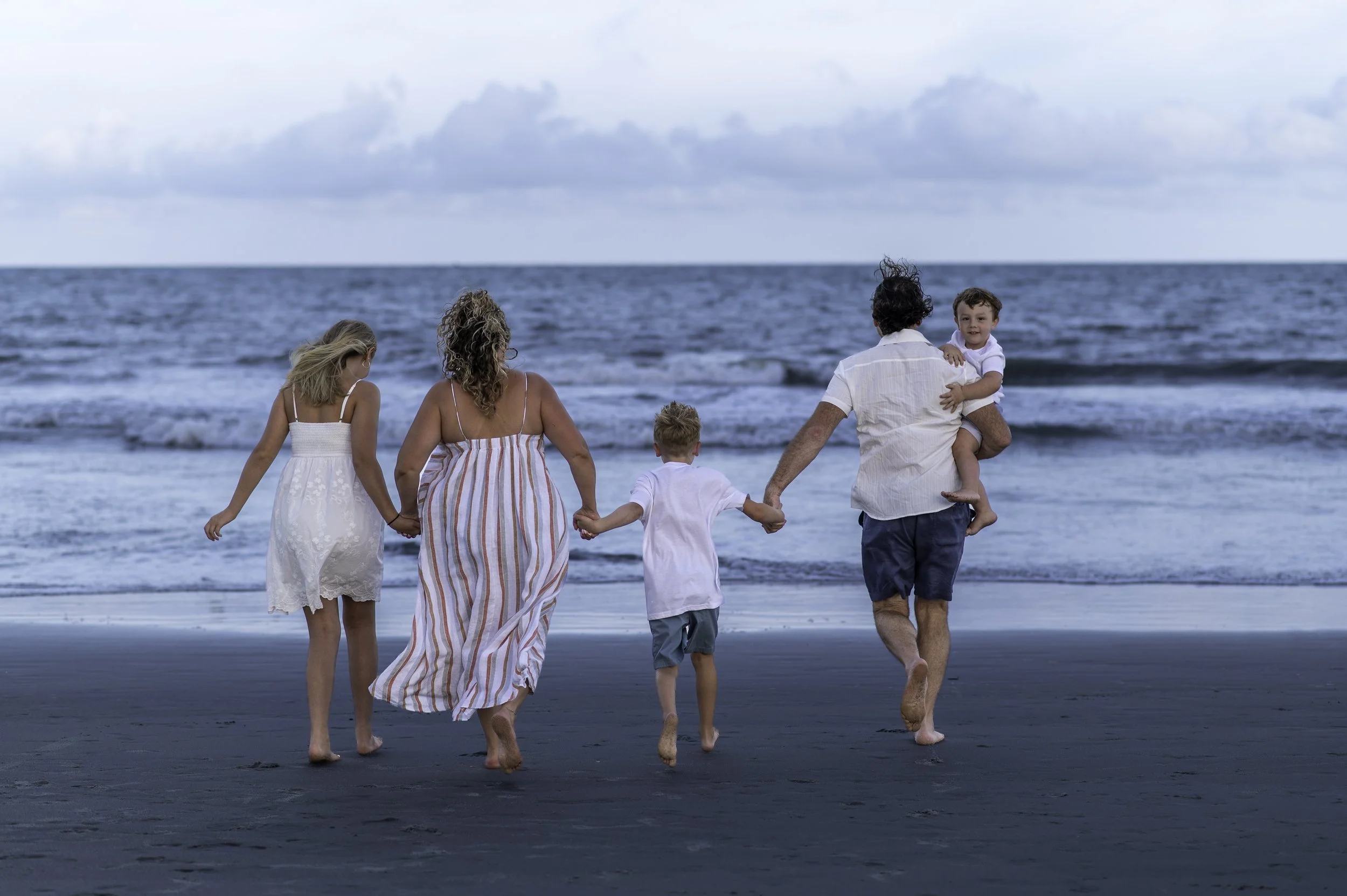 A family walking on the beach holding hands, with the ocean and cloudy sky in the background.