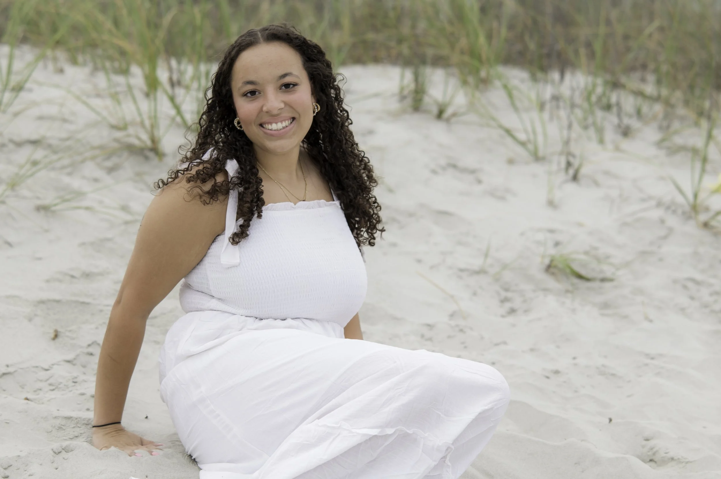 A young woman with curly hair, wearing a white sleeveless top and white skirt, sitting on the sandy beach with tall green grasses in the background, smiling at the camera.