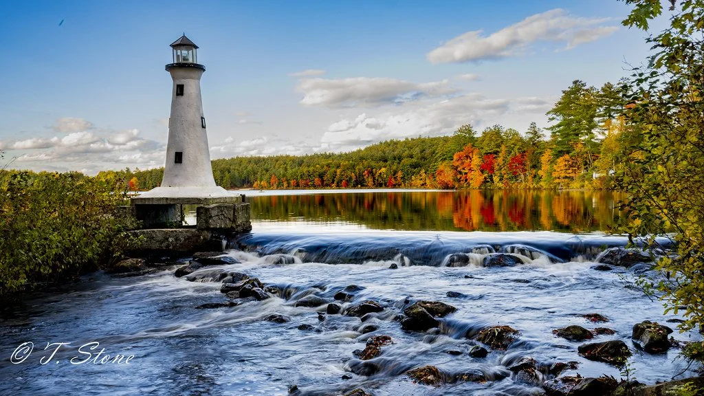 A lighthouse on a small rocky pier beside a flowing river, surrounded by trees with autumn foliage under a partly cloudy sky.