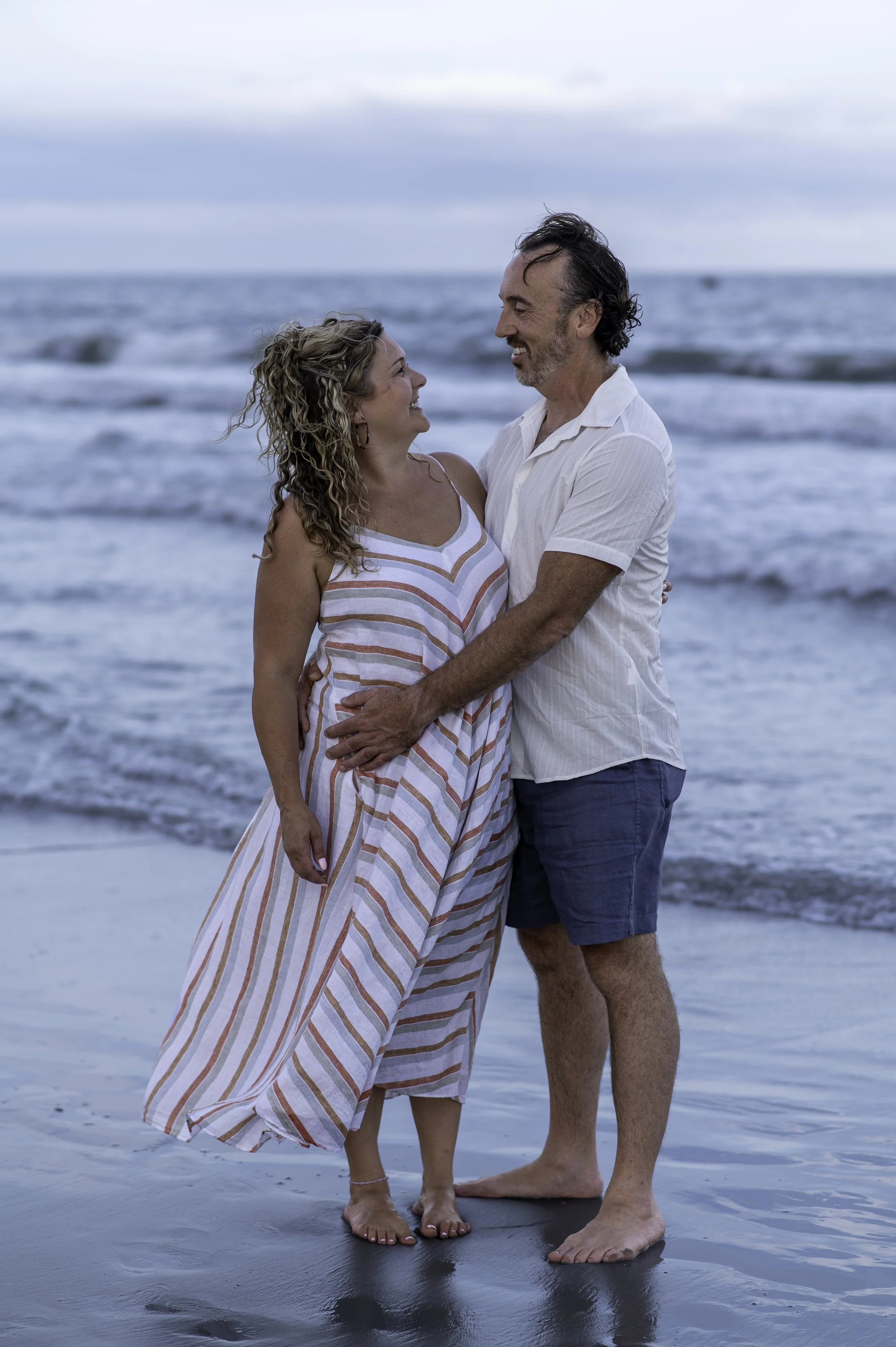 A couple standing on the beach at the ocean, smiling and looking at each other, holding each other, with the waves in the background.