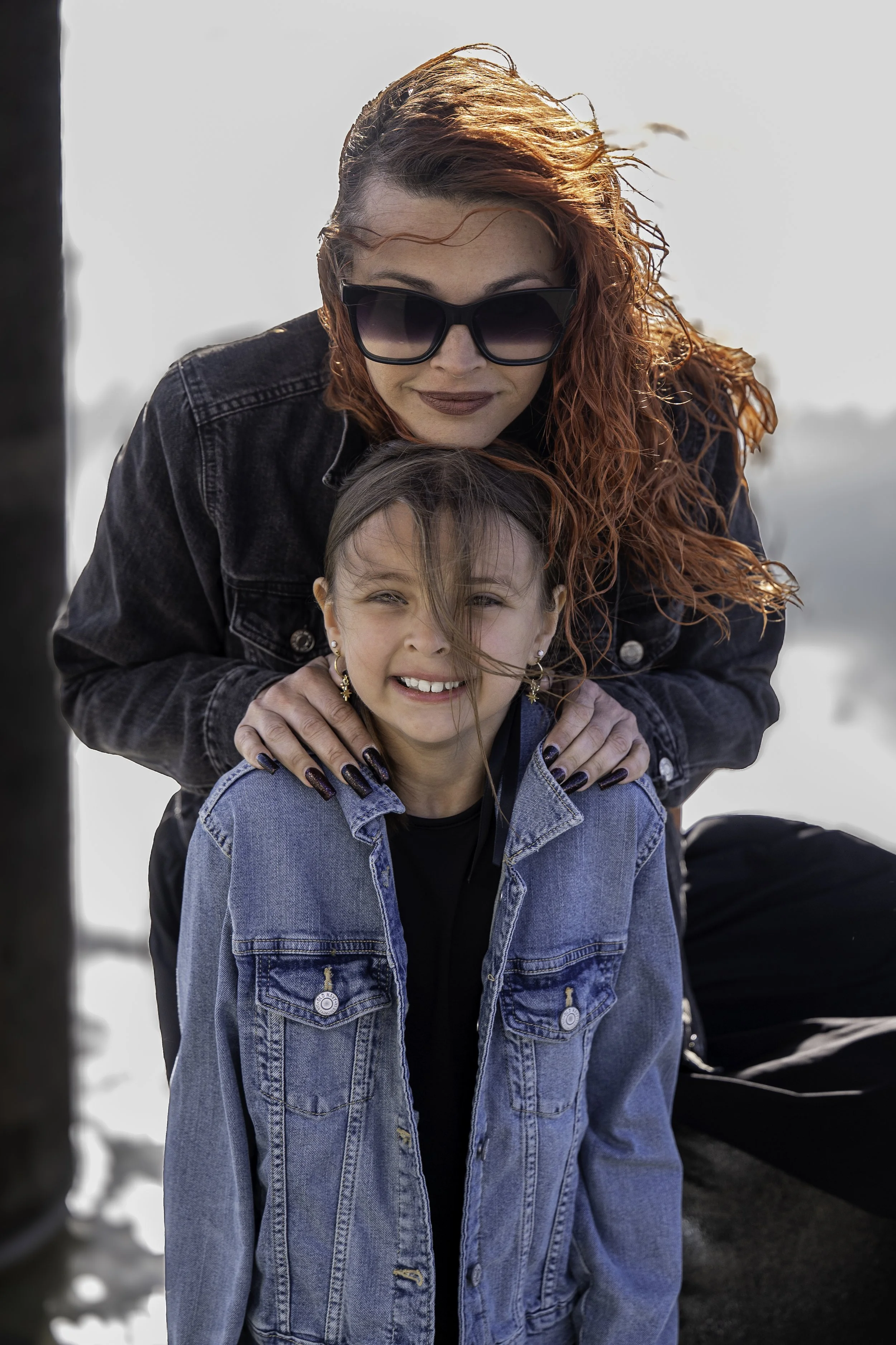 A woman with vibrant red hair wearing large sunglasses and a black jacket, smiling while hugging a young girl with straight brown hair, a denim jacket, and earrings, outdoors on a cloudy day.
