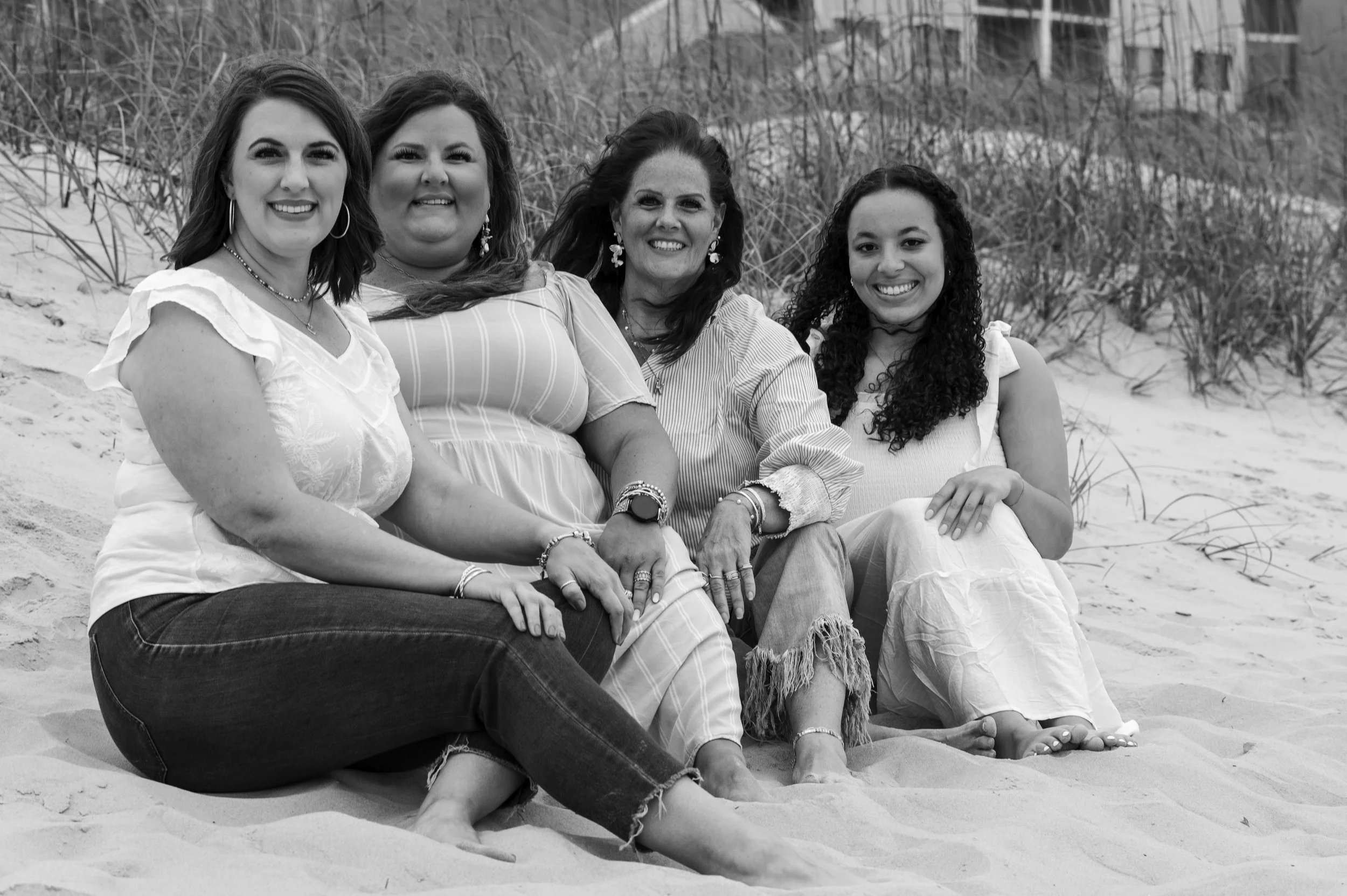 Group of four women sitting on a sandy beach with beach grass and a building in the background, smiling at the camera.