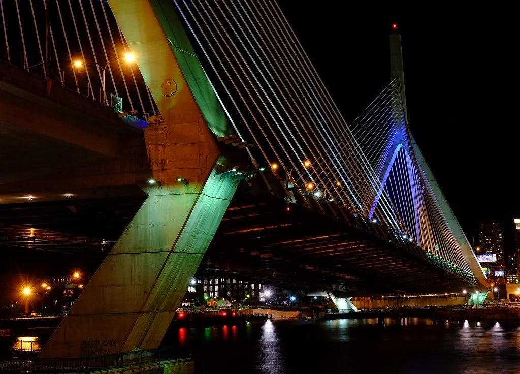 Night view of a modern cable-stayed bridge illuminated with green and blue lights, spanning over a river with city lights in the background.