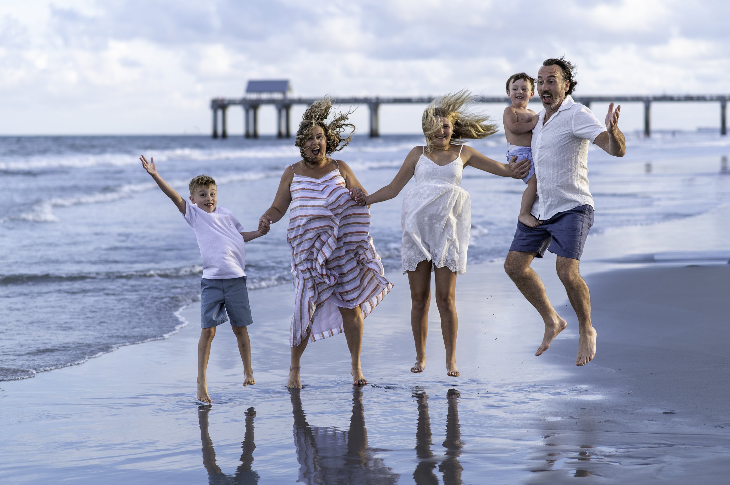 A family of five, including two children and three adults, holding hands and running along the beach near the water, with a pier in the background. They are smiling and enjoying a day at the beach.