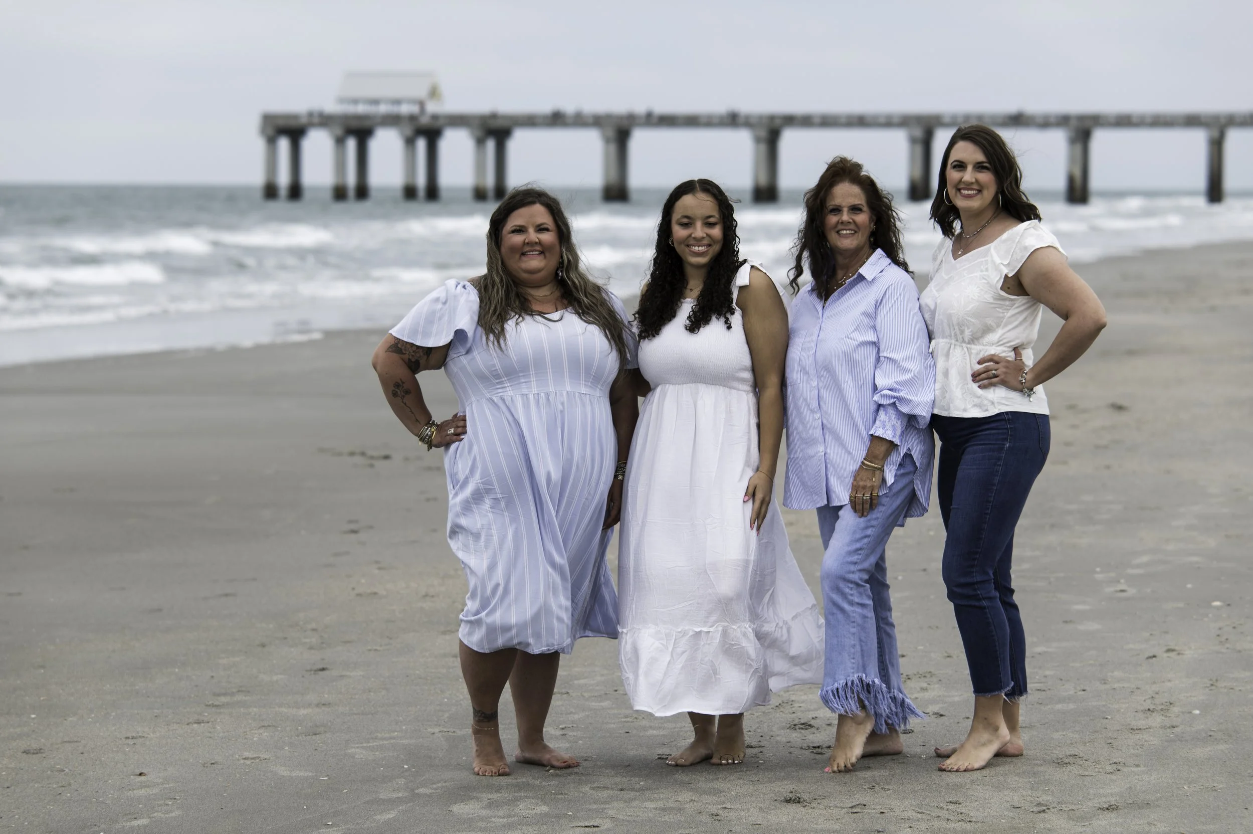 Four women standing barefoot on a sandy beach near the water, with a pier extending into the ocean in the background. They are smiling and dressed in casual white and blue dresses.