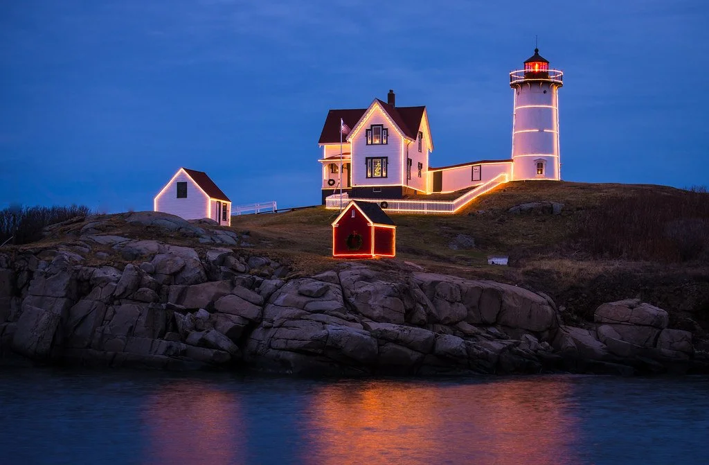 Lighthouse and houses decorated with colorful holiday lights on a rocky shoreline during sunset or dusk.