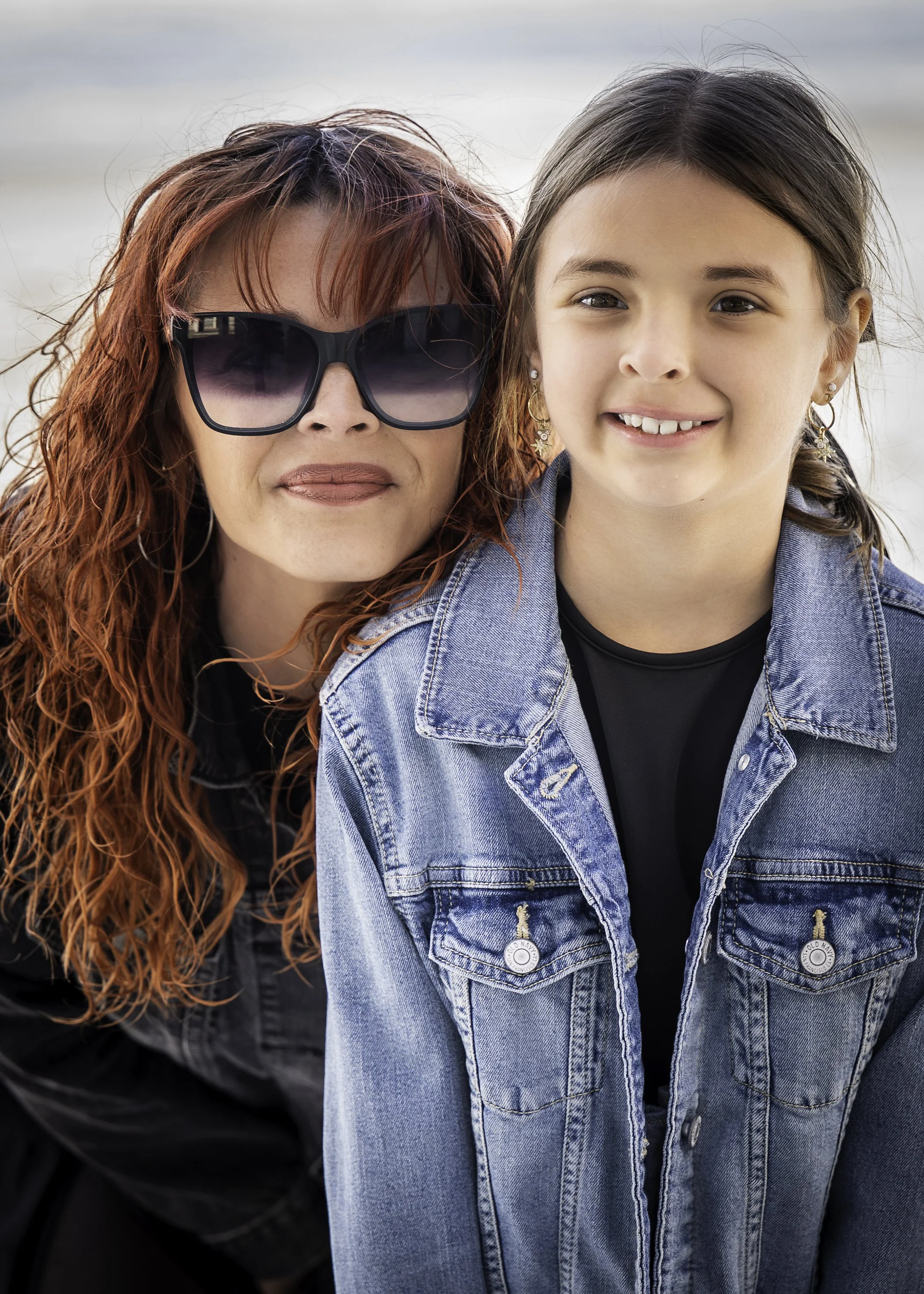 Close-up of a woman with red, curly hair wearing large black sunglasses and a girl with dark hair in a ponytail wearing gold jewelry and a denim jacket, both smiling outdoors.