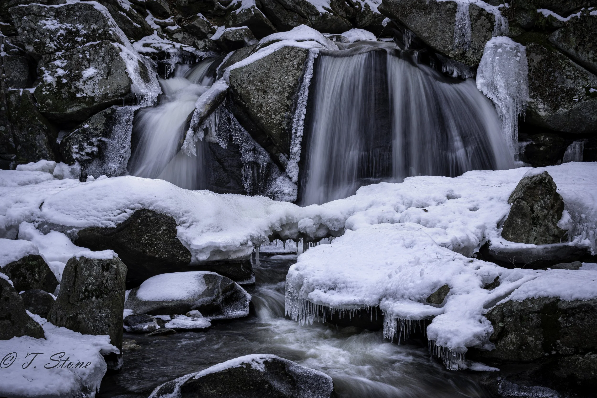 A small waterfall flowing over rocks in a snowy, icy landscape, with ice formations hanging from some rocks.