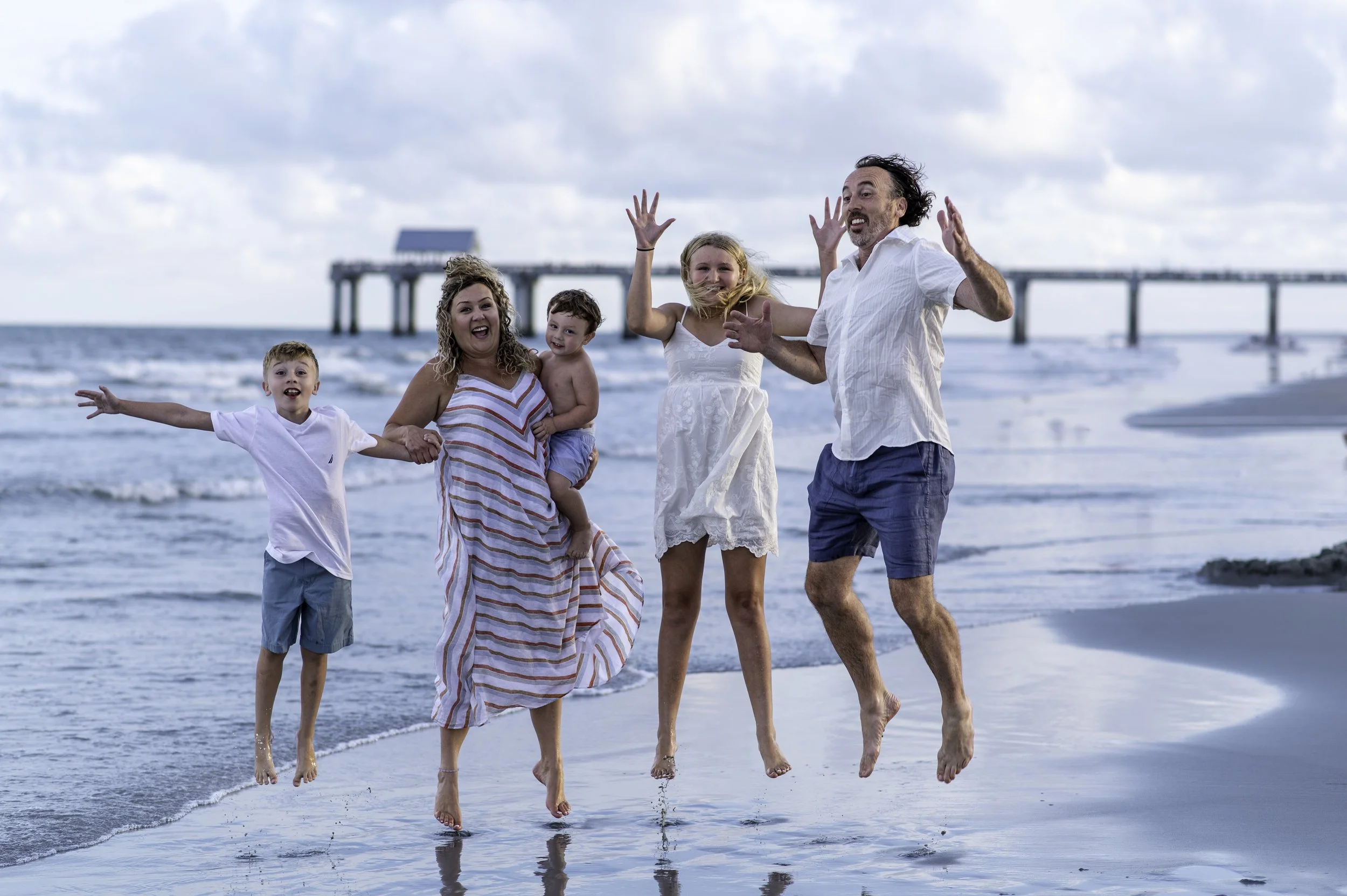 Family of six, including two young children, happily jumping and playing on the beach near the water with a pier in the background.