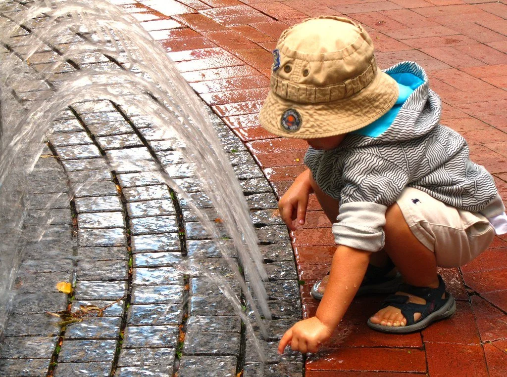 Child crouching by a water fountain touching the water with their finger, wearing a beige bucket hat, striped jacket, beige shorts, and sandals, on a wet brick sidewalk.