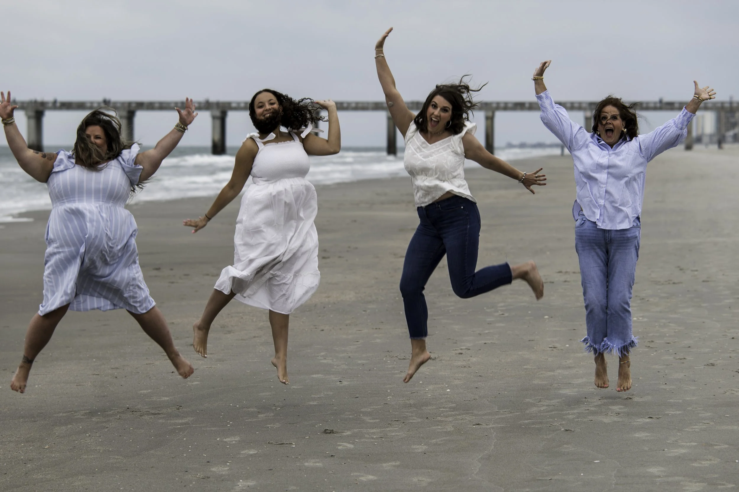Four women jumping and dancing on a beach, with a pier in the background, enjoying a cloudy day.