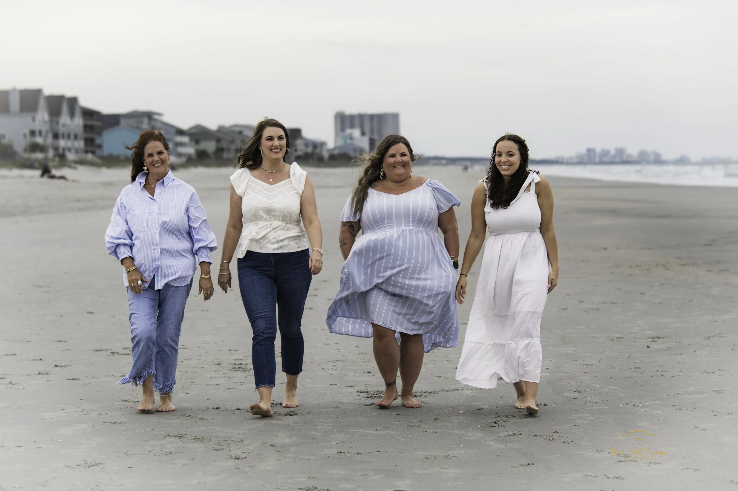 Four women walking barefoot on a beach, smiling and enjoying a cloudy day. In the background, there are beach houses and a city skyline.