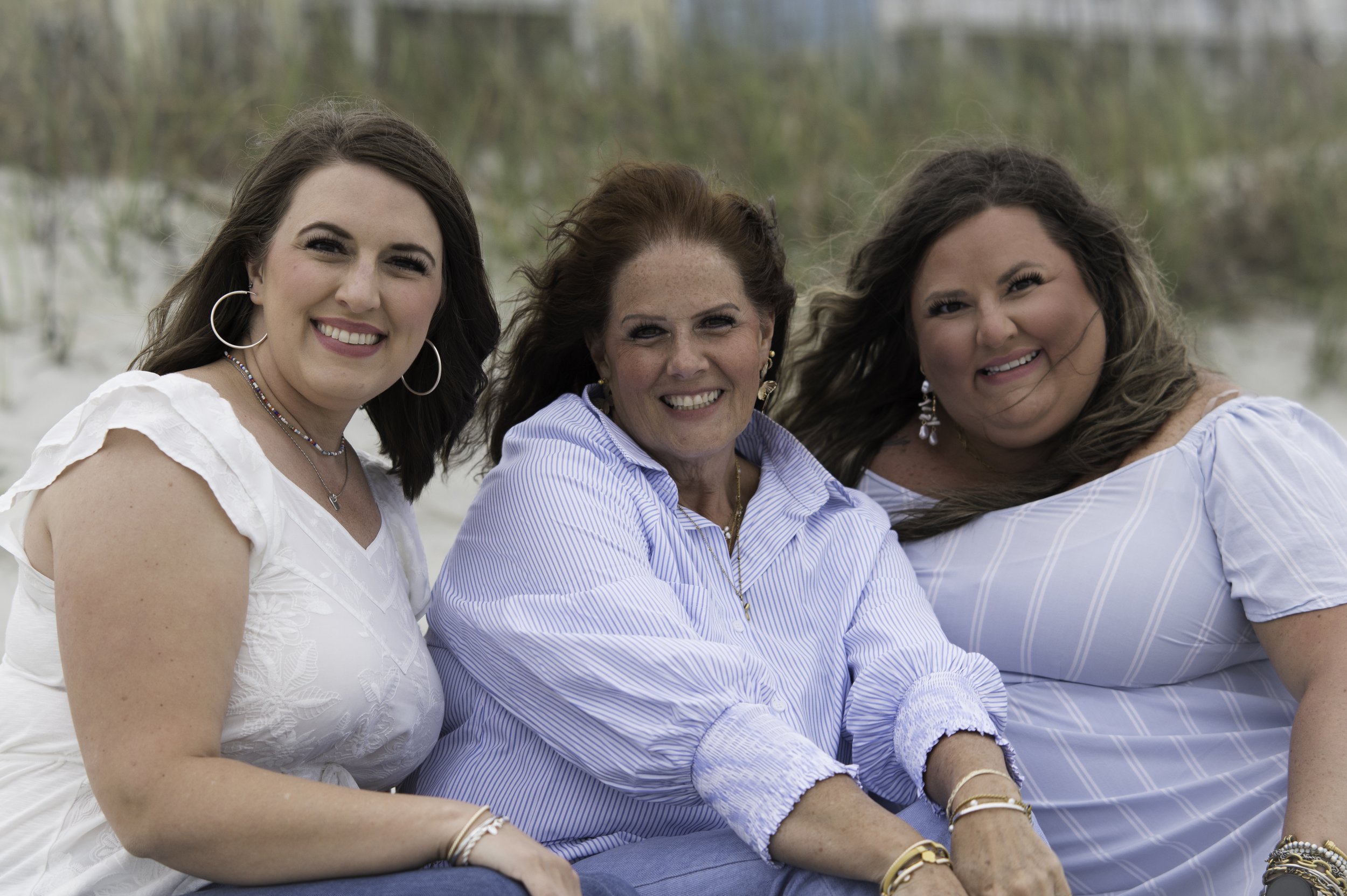 Three women sitting outdoors with blurred trees in the background, smiling and looking at the camera.