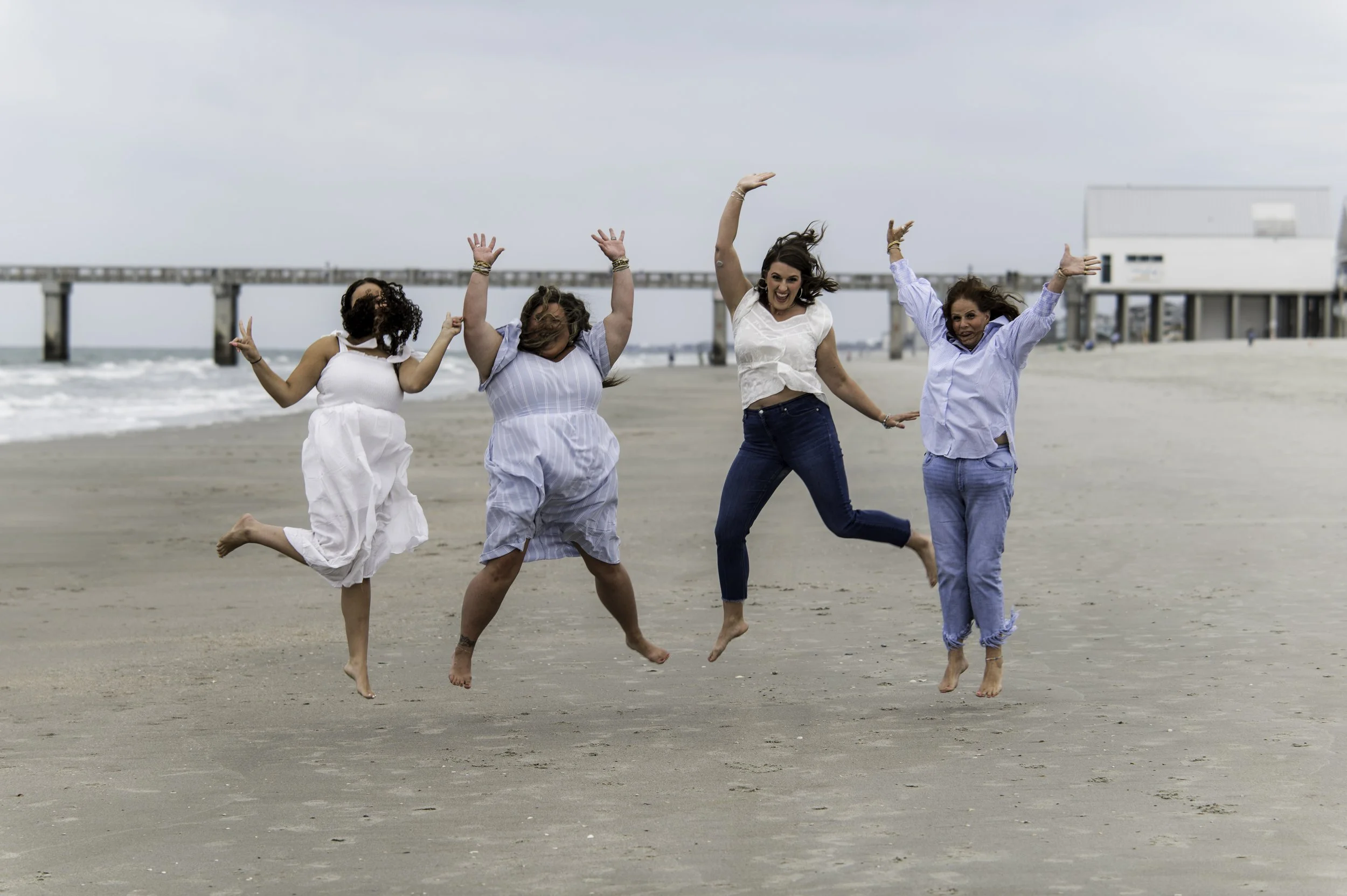 Four women celebrating and jumping on a beach with a pier in the background.