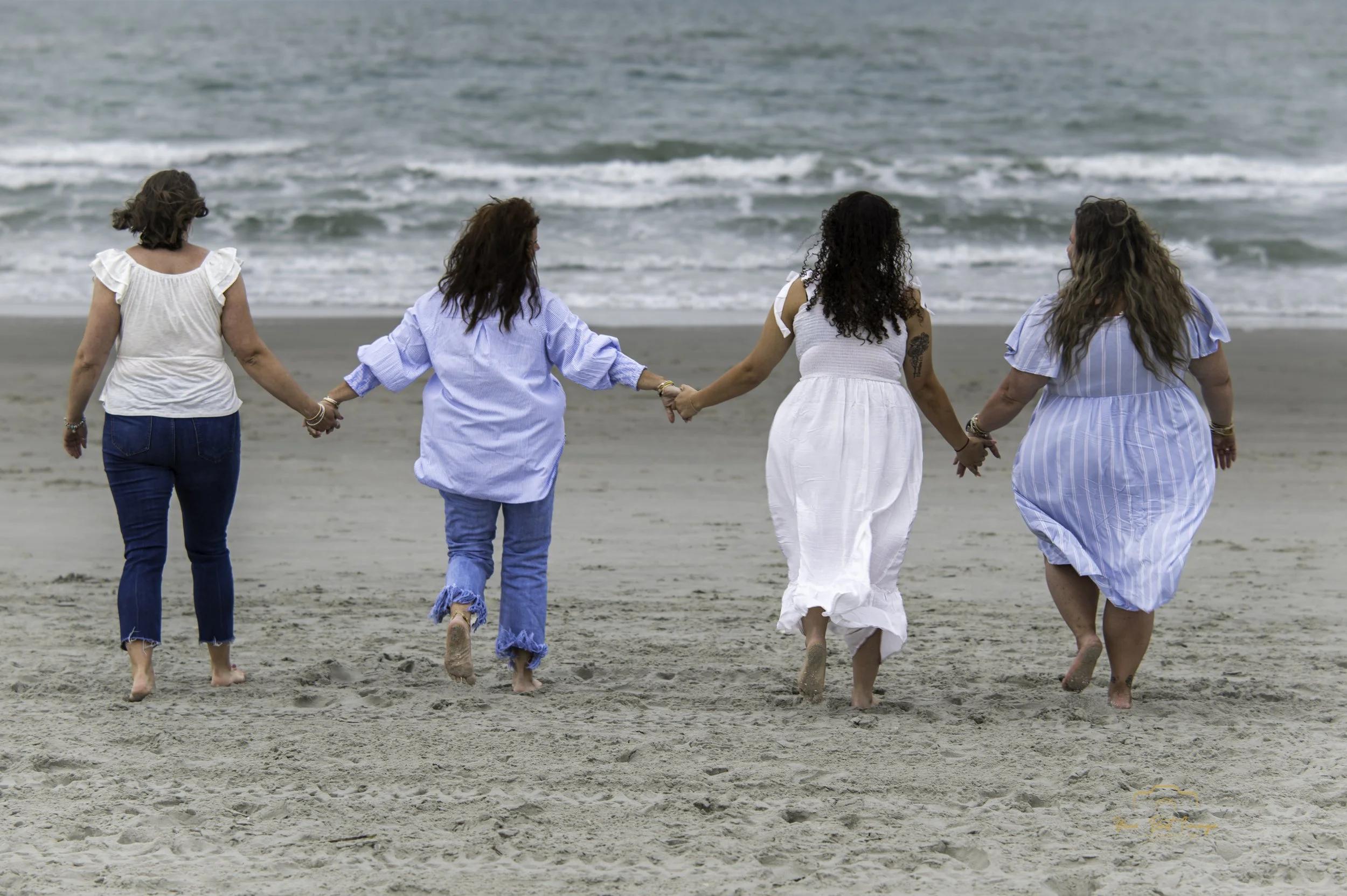 Four women walking hand-in-hand on a beach. They are dressed in casual summer clothes and barefoot, with ocean waves visible in the background.
