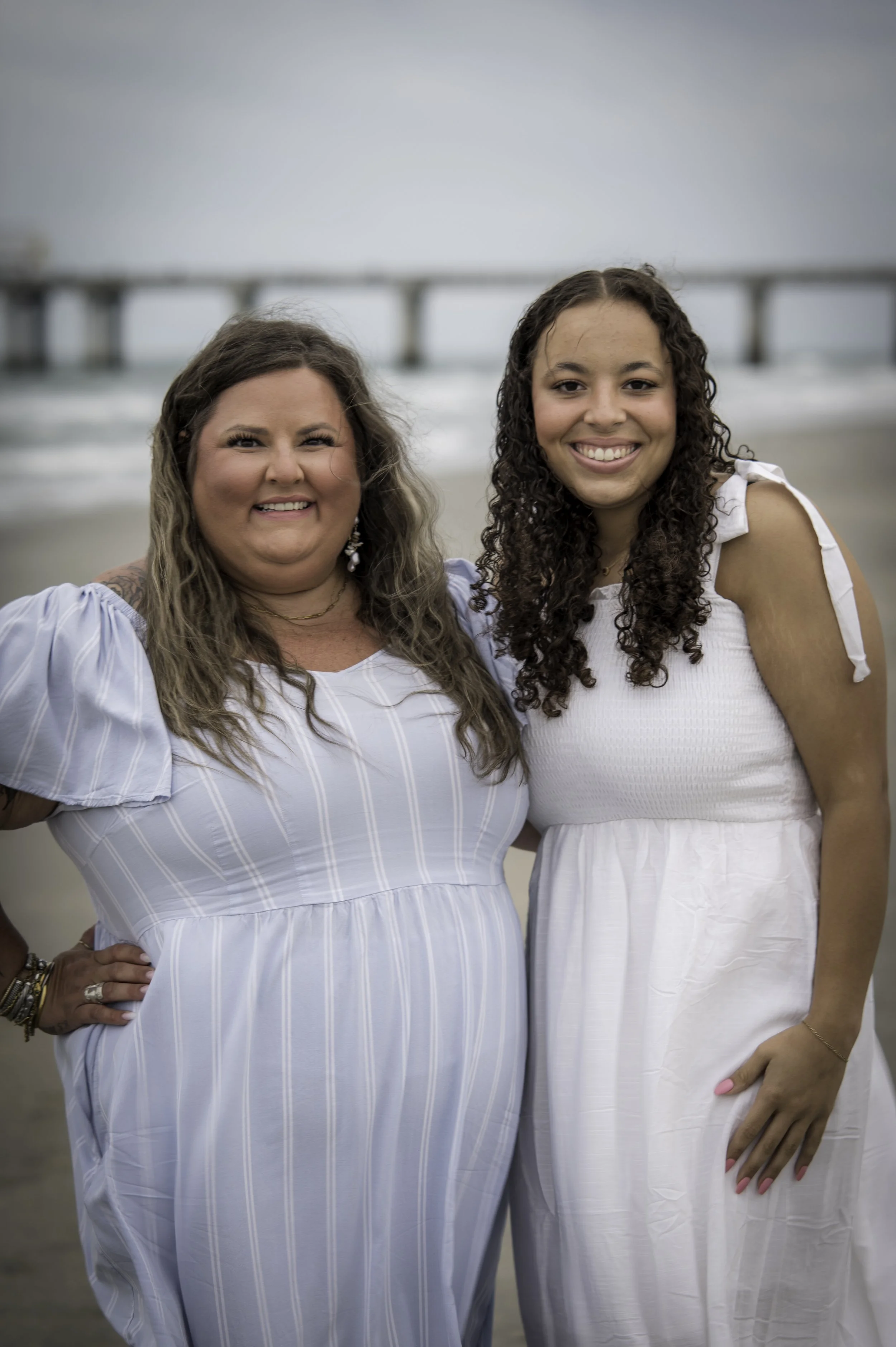 Two women smiling at the beach with a pier in the background. One woman has long, wavy, light brown hair and is wearing a light blue striped dress. The other woman has long, curly, dark brown hair and is dressed in a white sleeveless dress.