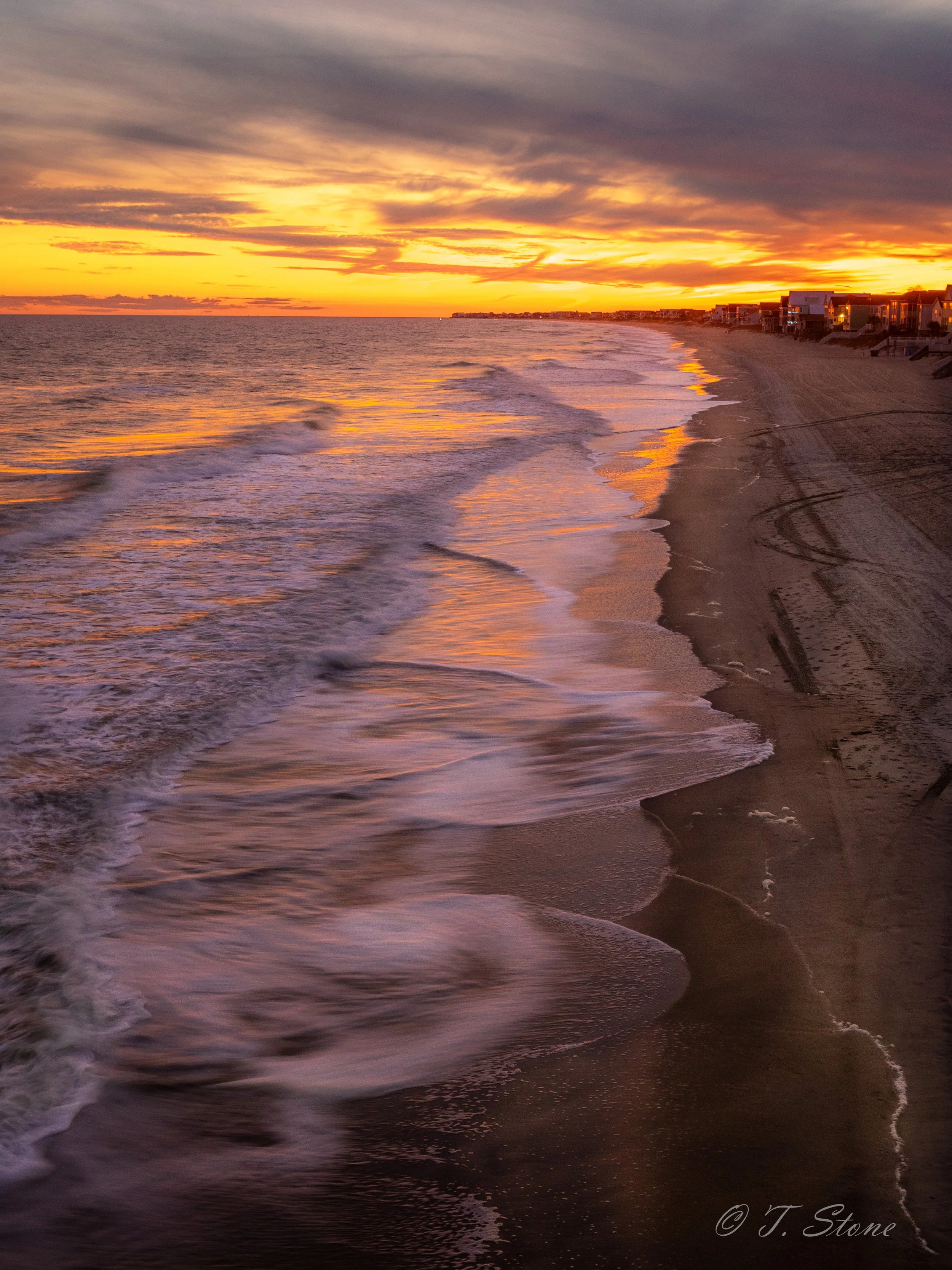 Sunset over the ocean with waves crashing onto a sandy beach, houses along the shoreline, and colorful clouds in the sky.