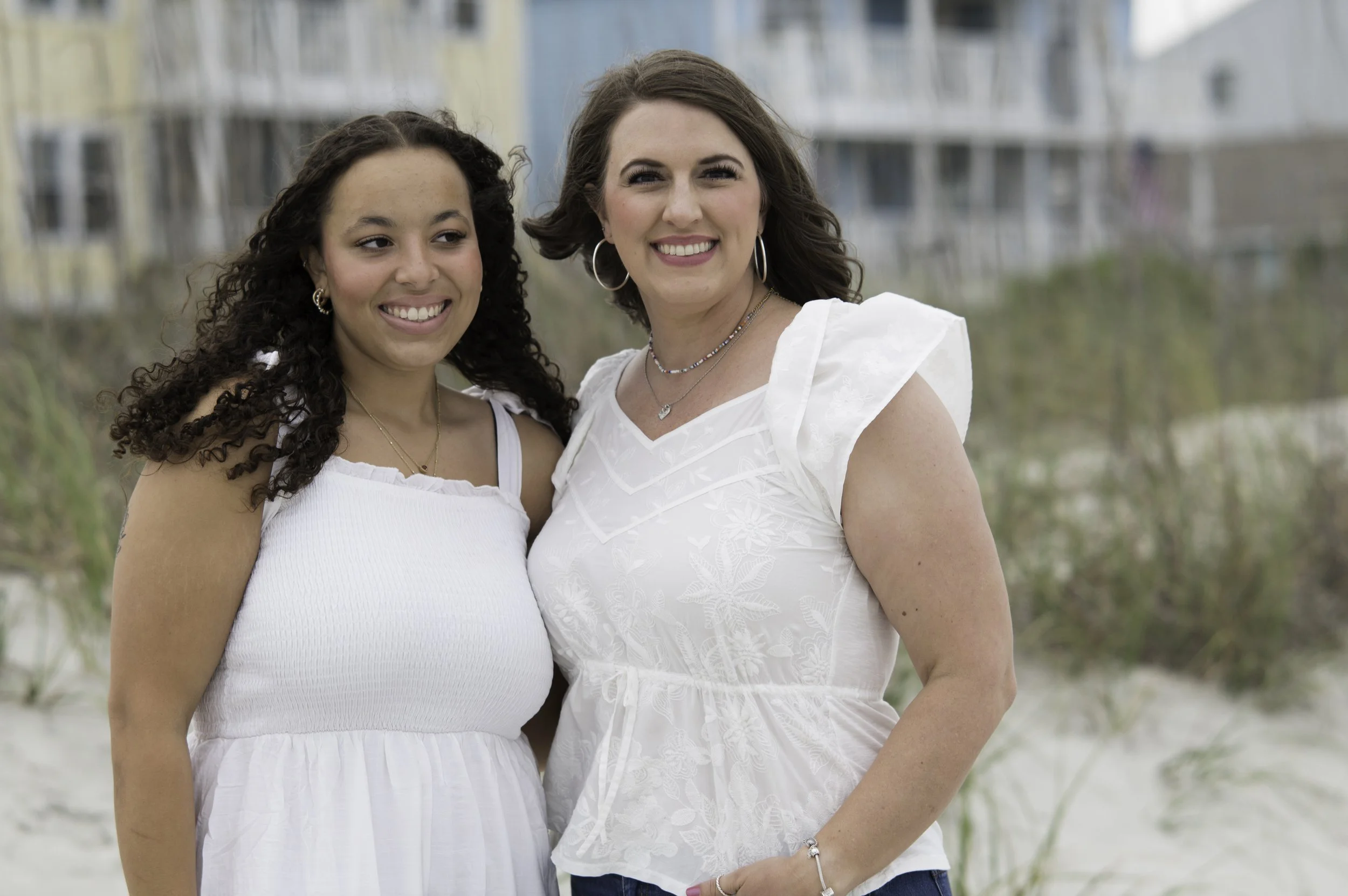 Two women smiling outdoors with a beach background, wearing white tops and jewelry.