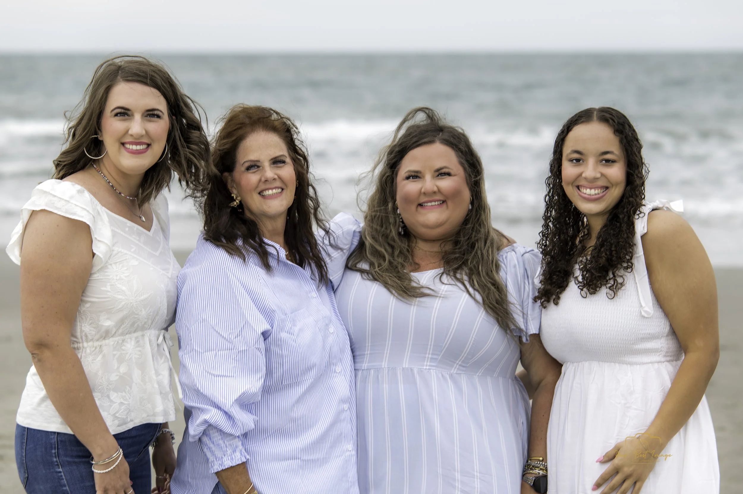 Group of four women smiling and posing on a beach with the ocean in the background.