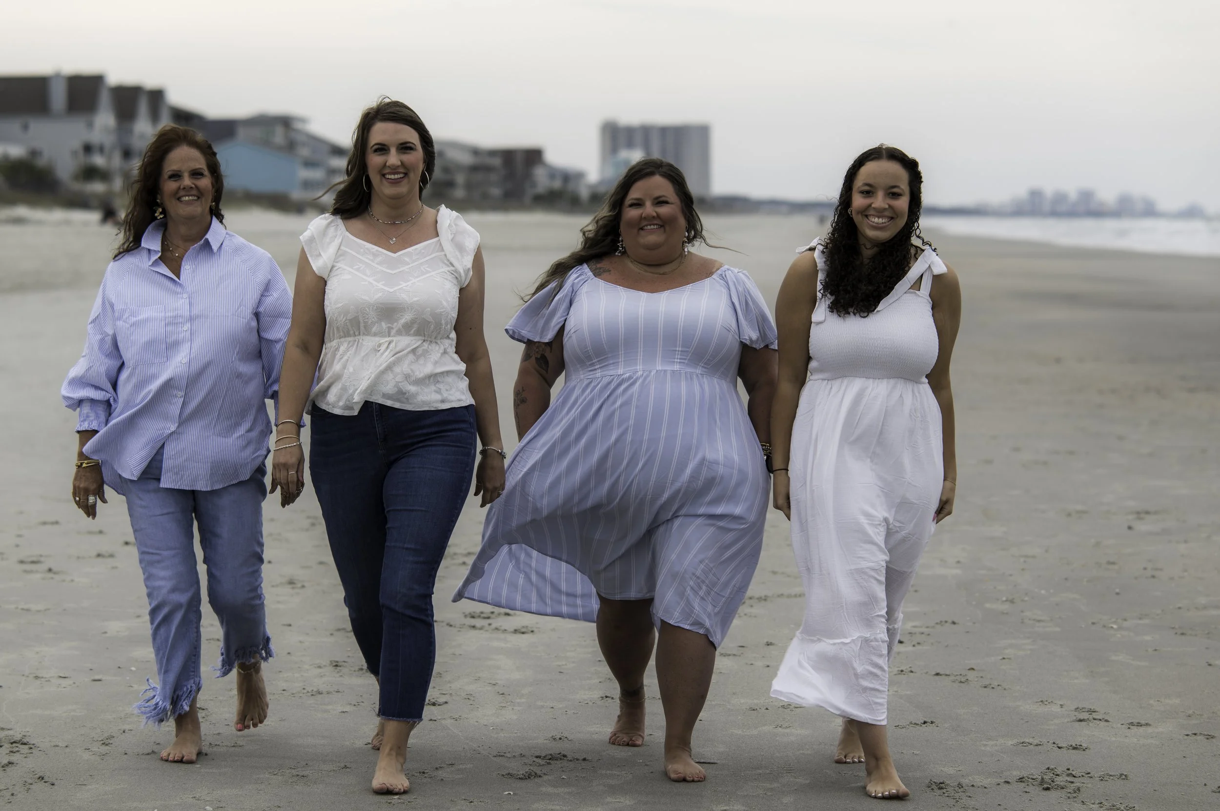 Four women walking on the beach holding hands, smiling, with houses and buildings in the background.