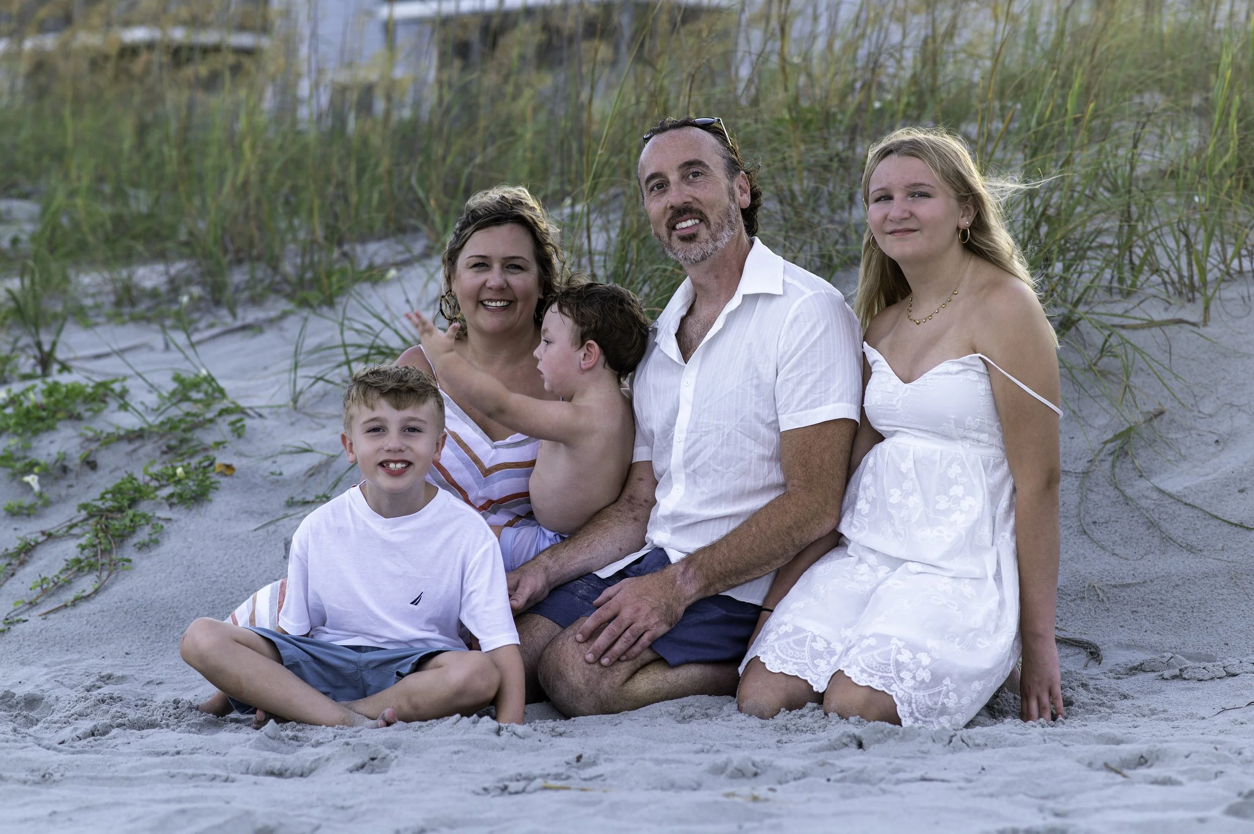 A family of six at the beach, sitting on the sand surrounded by grassy dunes and looking at the camera.