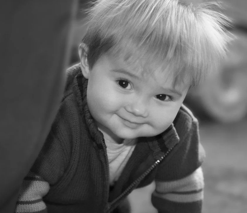 A smiling young child with light hair, wearing a striped sweater, looking up at the camera in black and white.