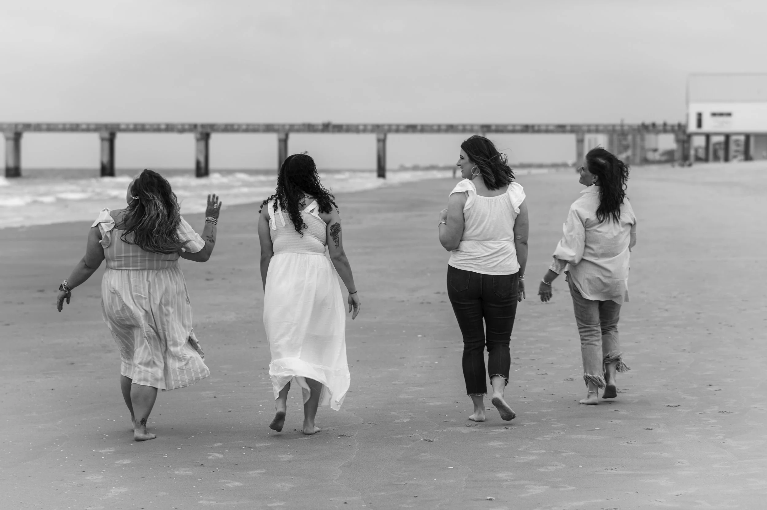 Four women walking on the beach near the ocean with a pier in the background, all dressed in light-colored clothing, some with long hair, in a black-and-white photo.