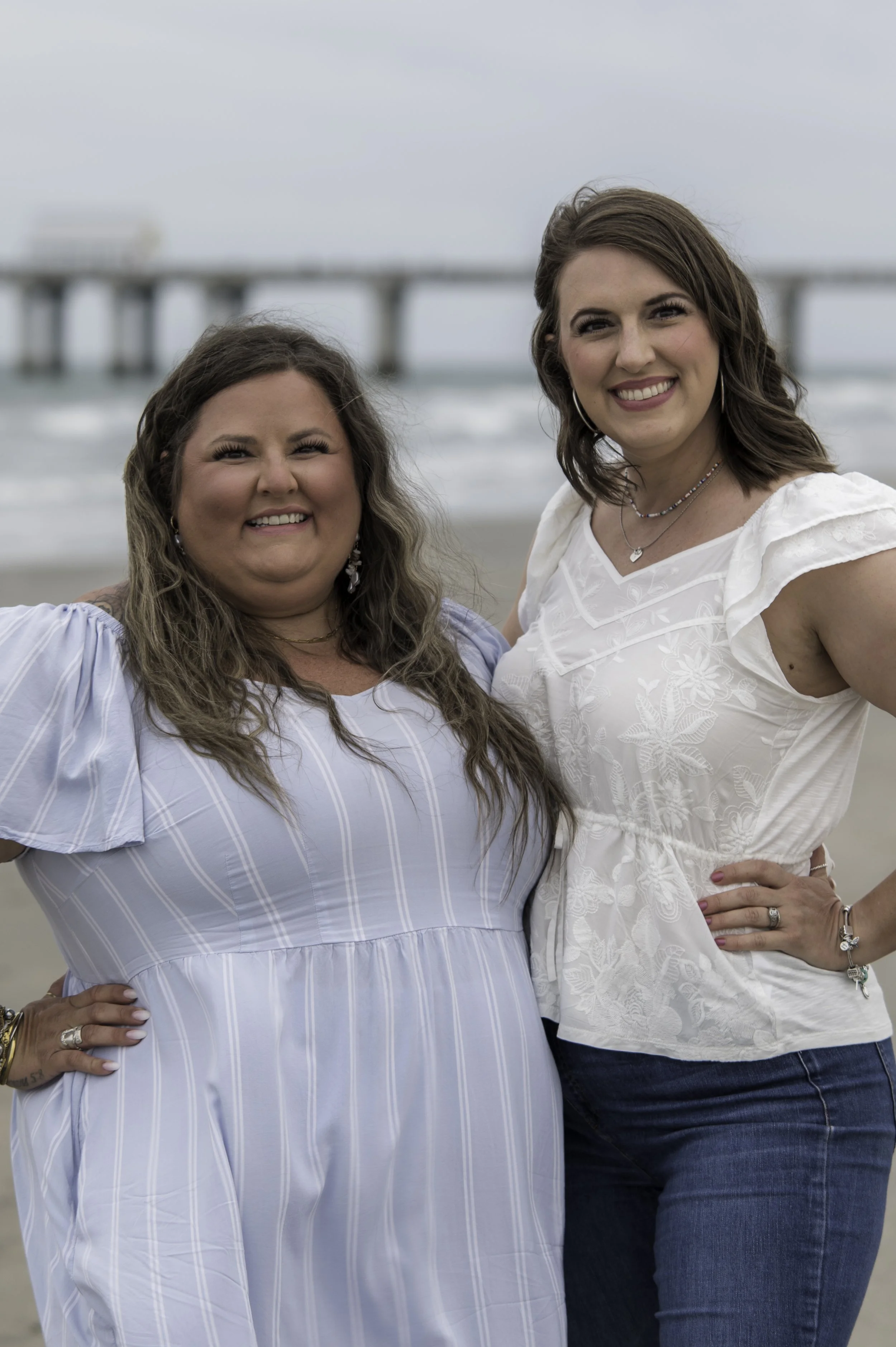Two women smiling on a beach with a pier in the background, one with long curly hair in a light blue dress, and the other with straight brown hair in a white top and jeans.