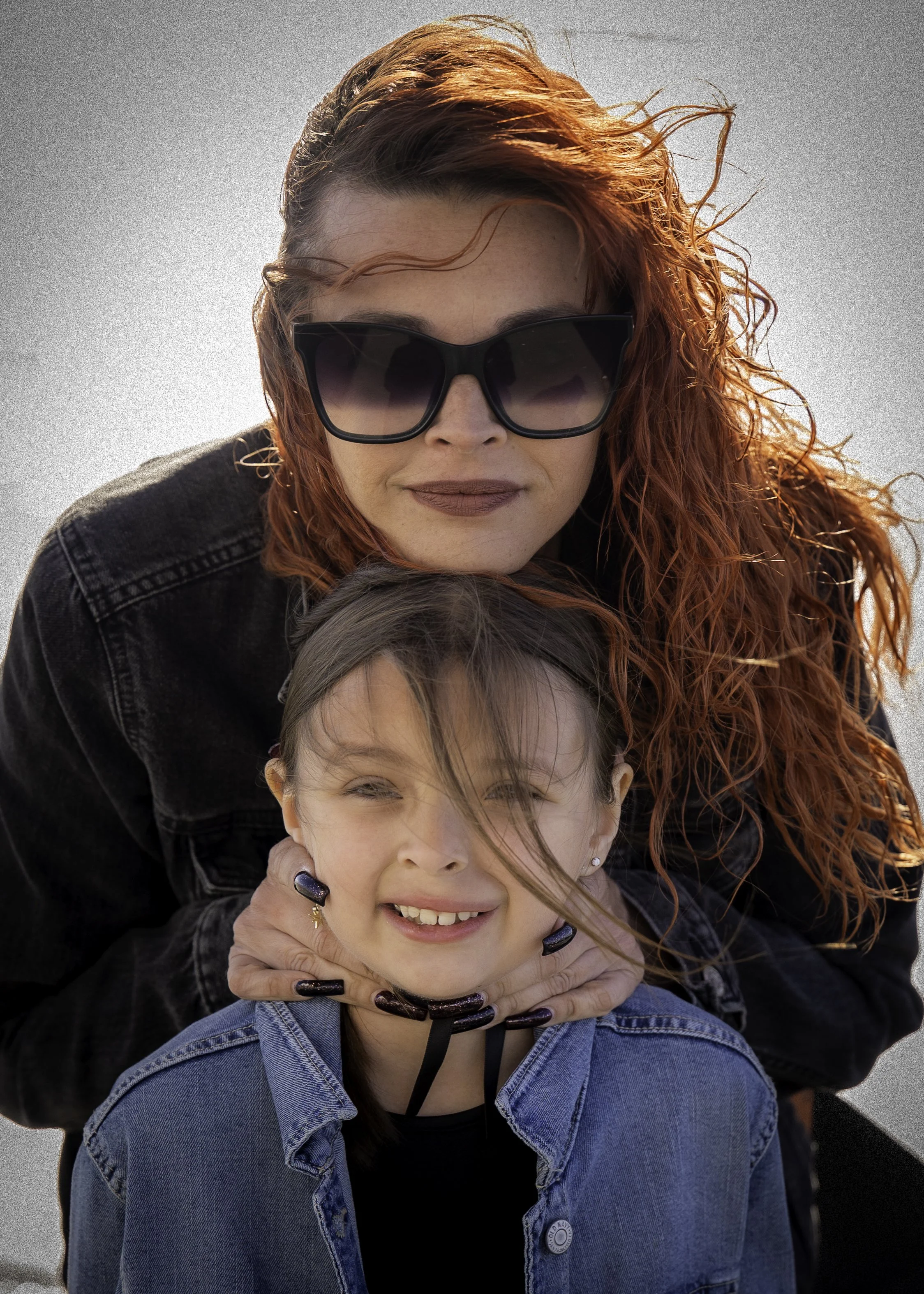 A woman with curly red hair wearing black sunglasses hugging a smiling girl with dark hair, a denim jacket, and ear piercings.