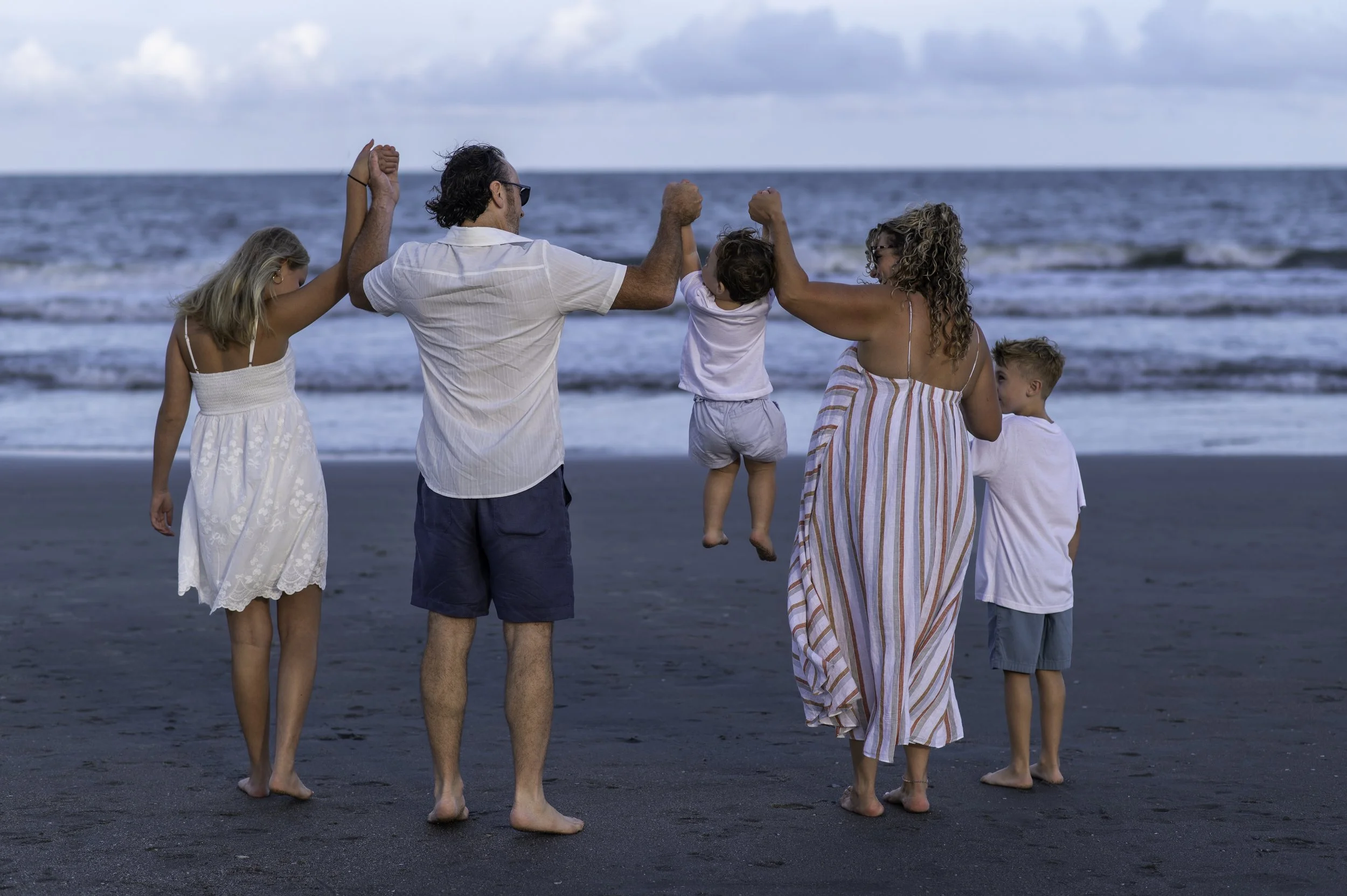 A family of five walking and celebrating together on the beach near the ocean, with the parents holding their children between them, all smiling and enjoying the moment.