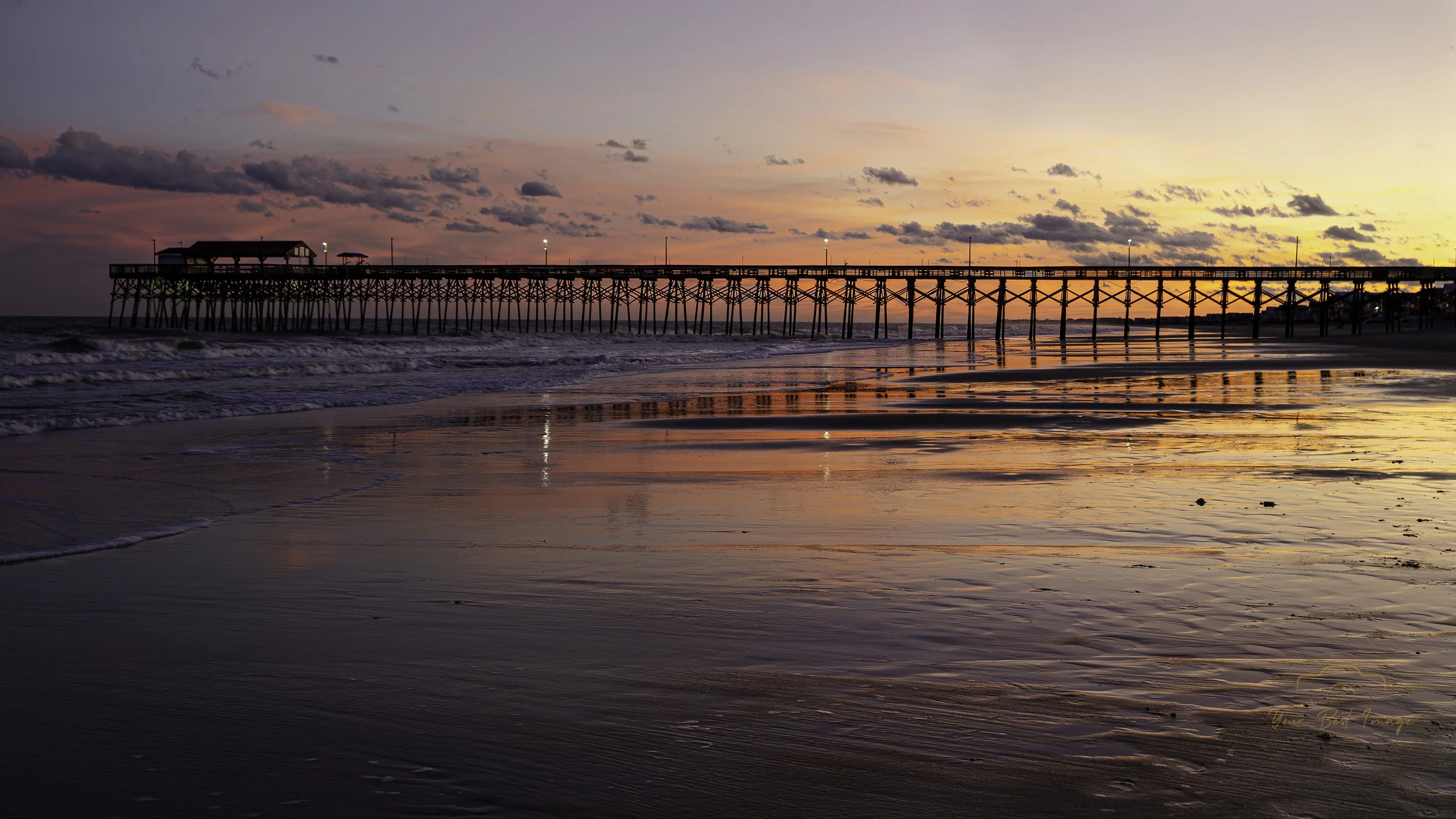 Sunset over a beach with a long pier extending into the water, calm waves lapping at the shore, and a few clouds in the sky.
