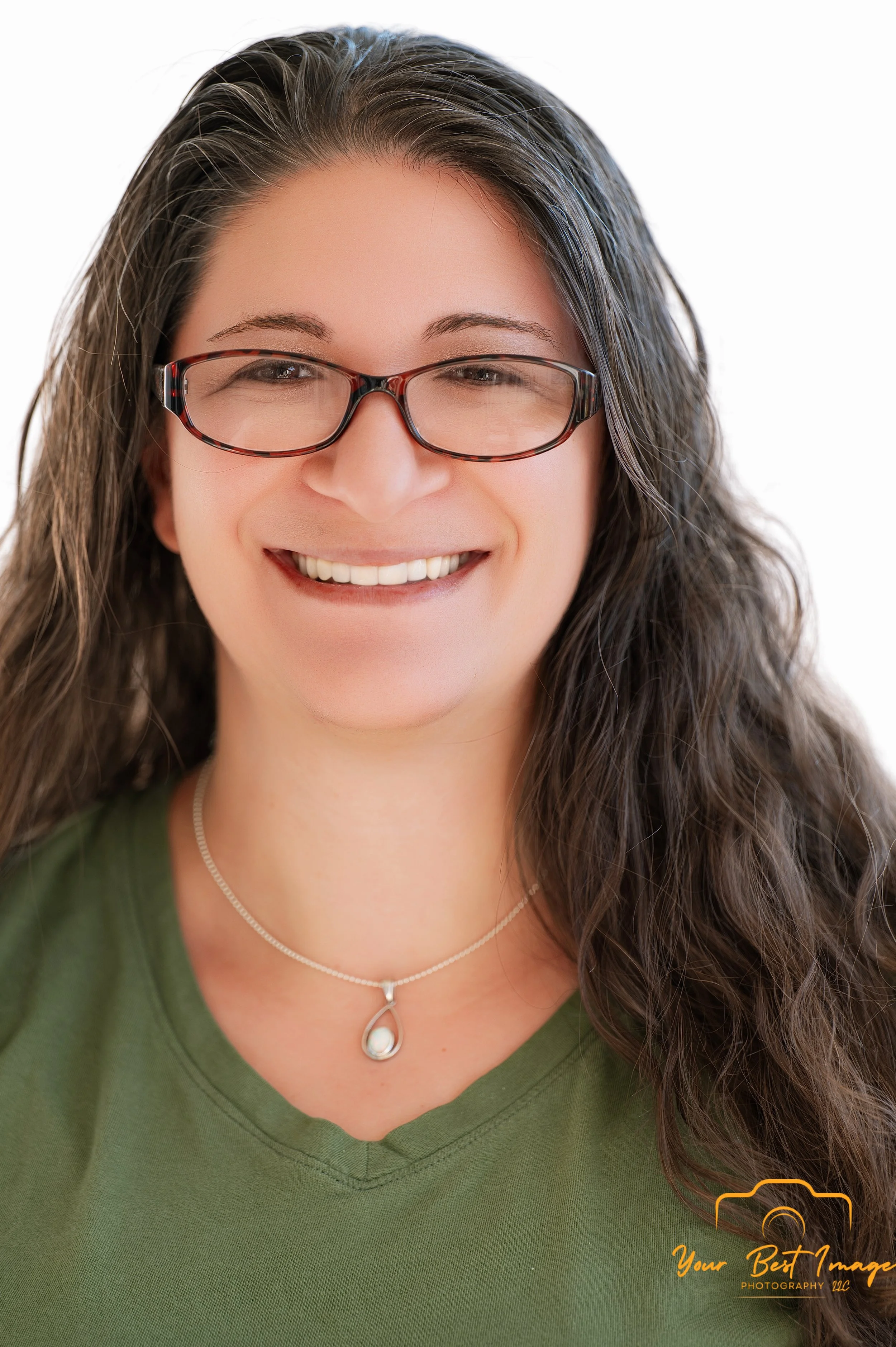 A woman with long brown hair and glasses, smiling, wearing a green shirt and a pearl necklace with a teardrop pendant.