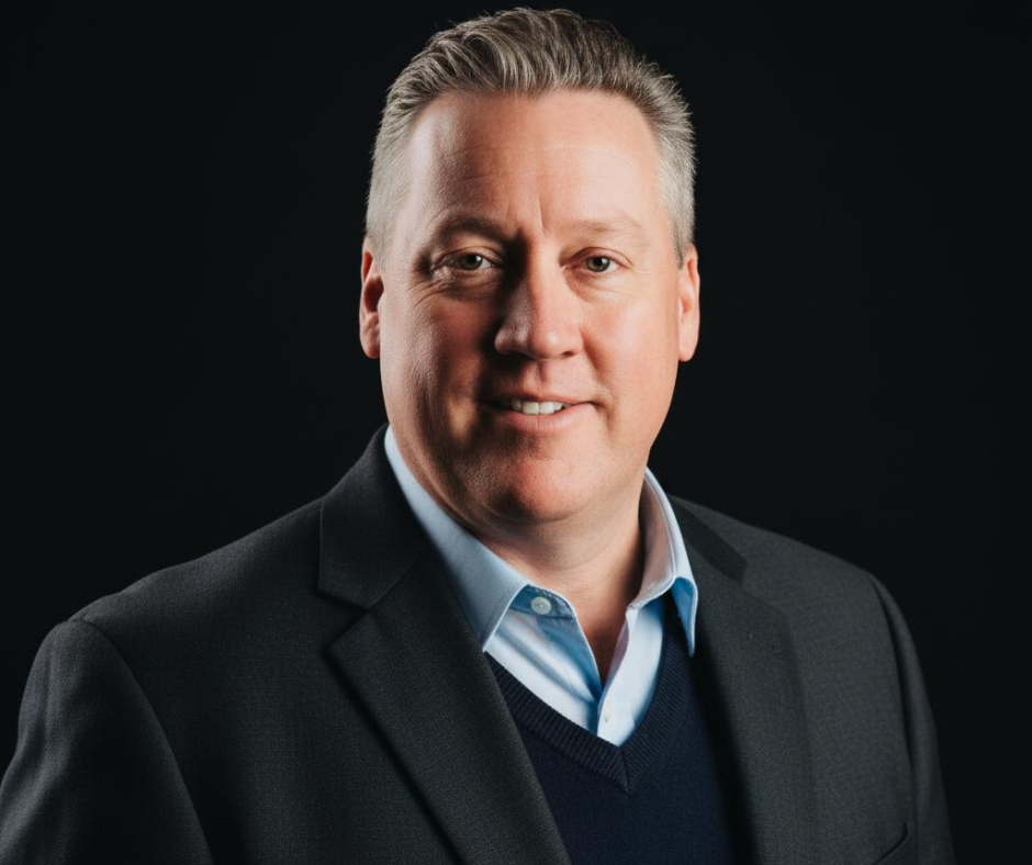 A professional headshot of a middle-aged man in a suit with a dark background.