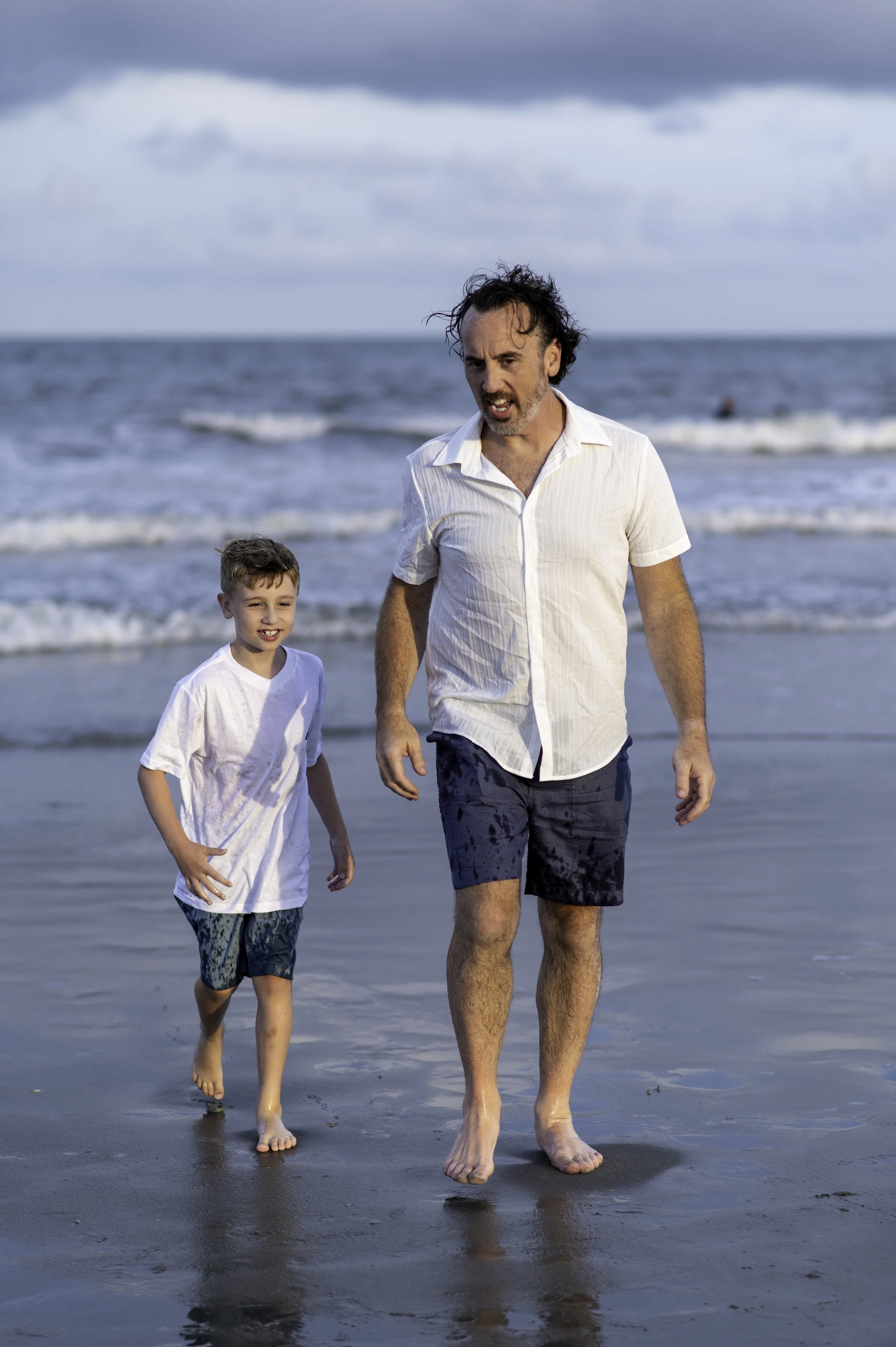 A man and a young boy walking barefoot on the wet sand at the beach with the ocean and cloudy sky in the background.