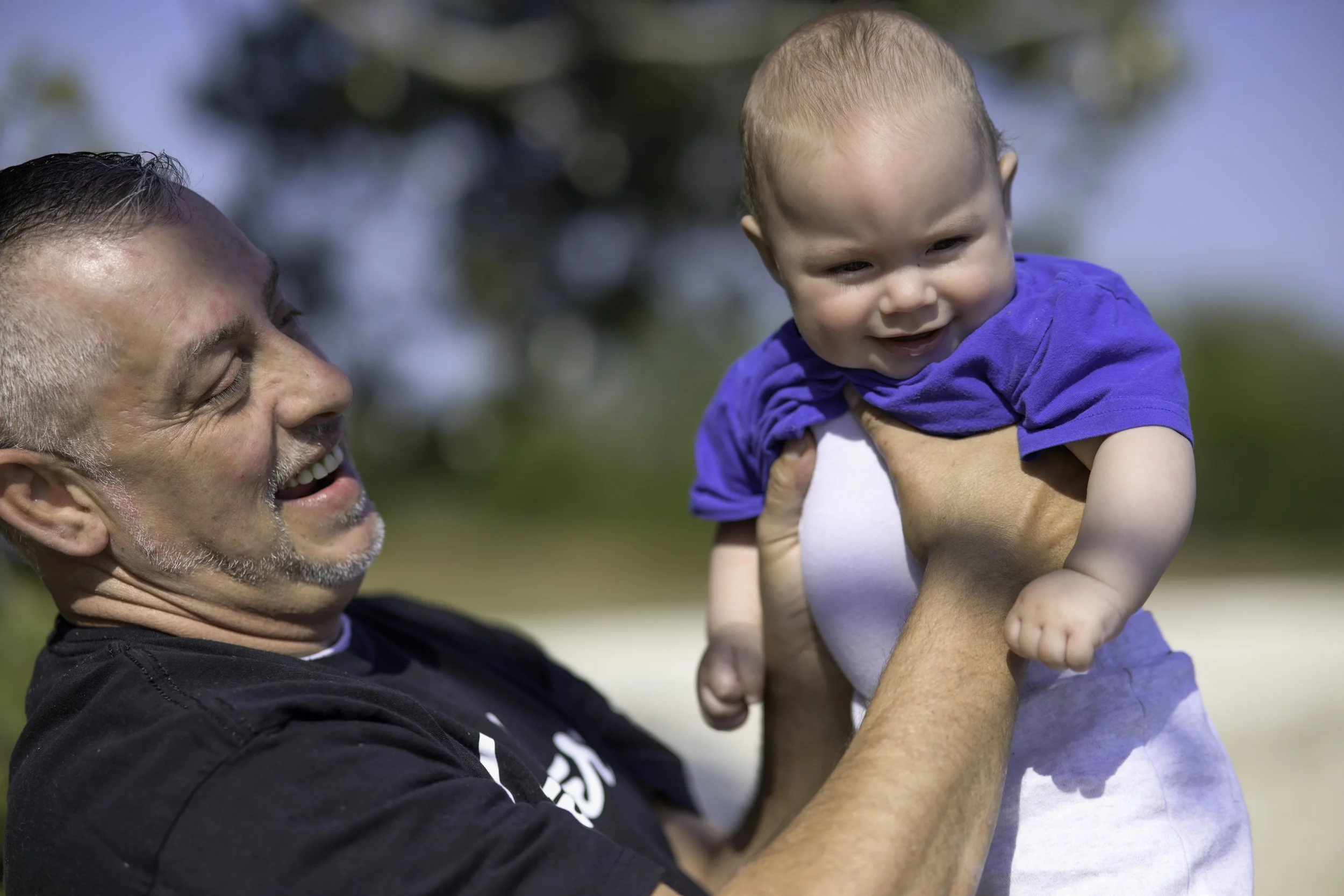 An older man with short dark hair and a beard is smiling and holding up a young toddler with short blonde hair and a big smile. The man is wearing a black shirt, and the child is wearing a blue shirt. They are outdoors with a background of blurred tr